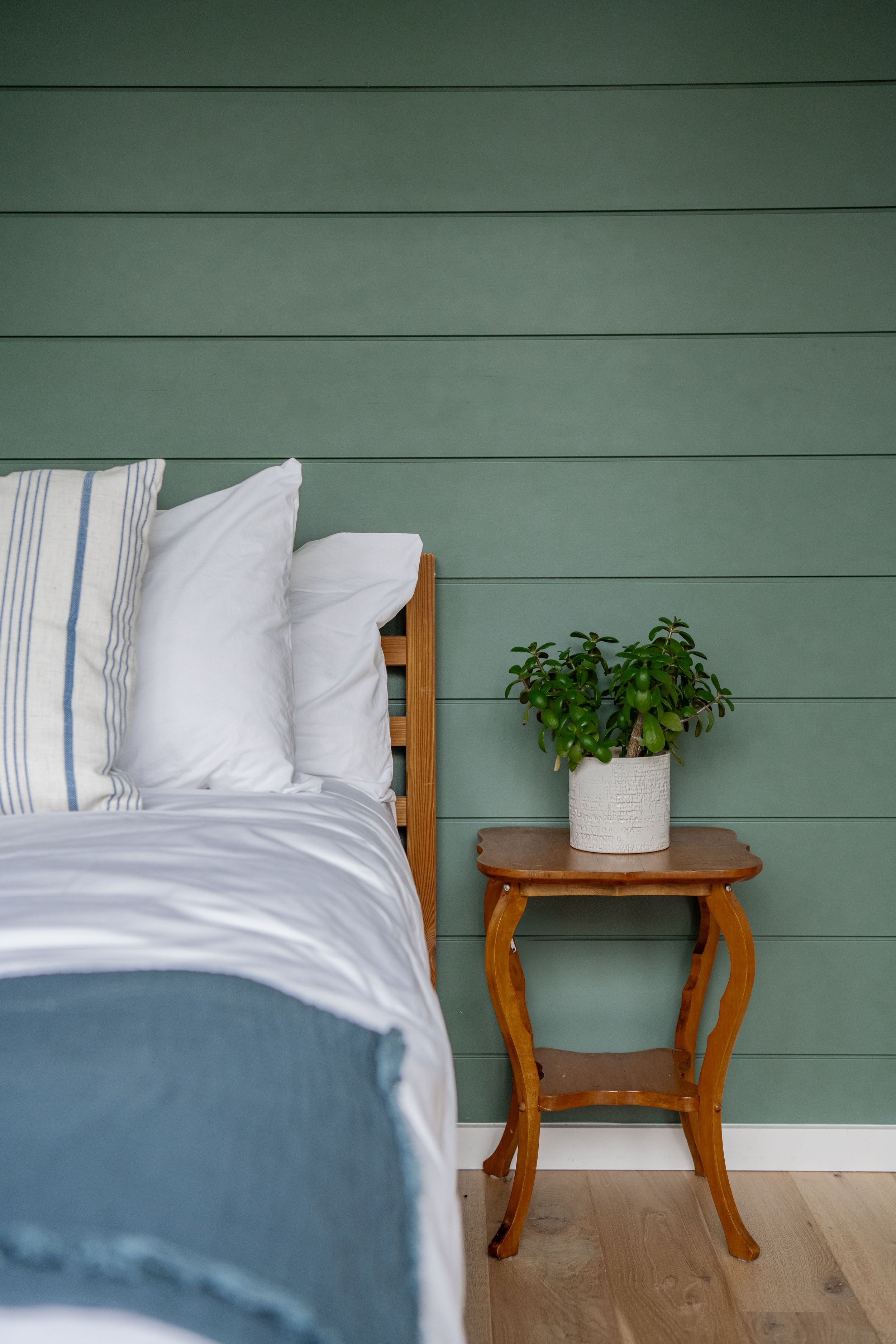 Cozy bedroom detail with white linens, sage green walls and wooden side table.