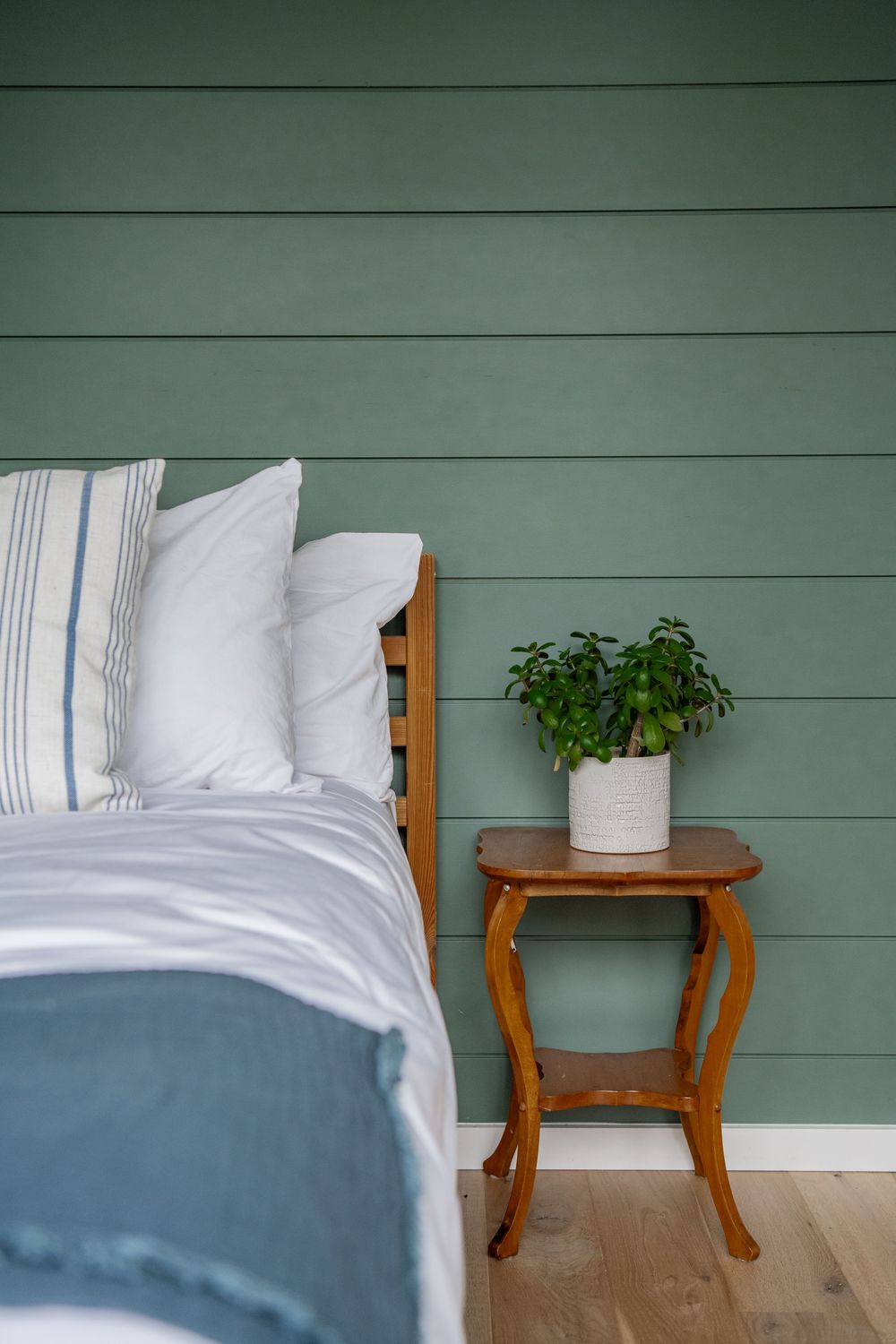 Cozy bedroom detail with white linens, sage green walls and wooden side table.