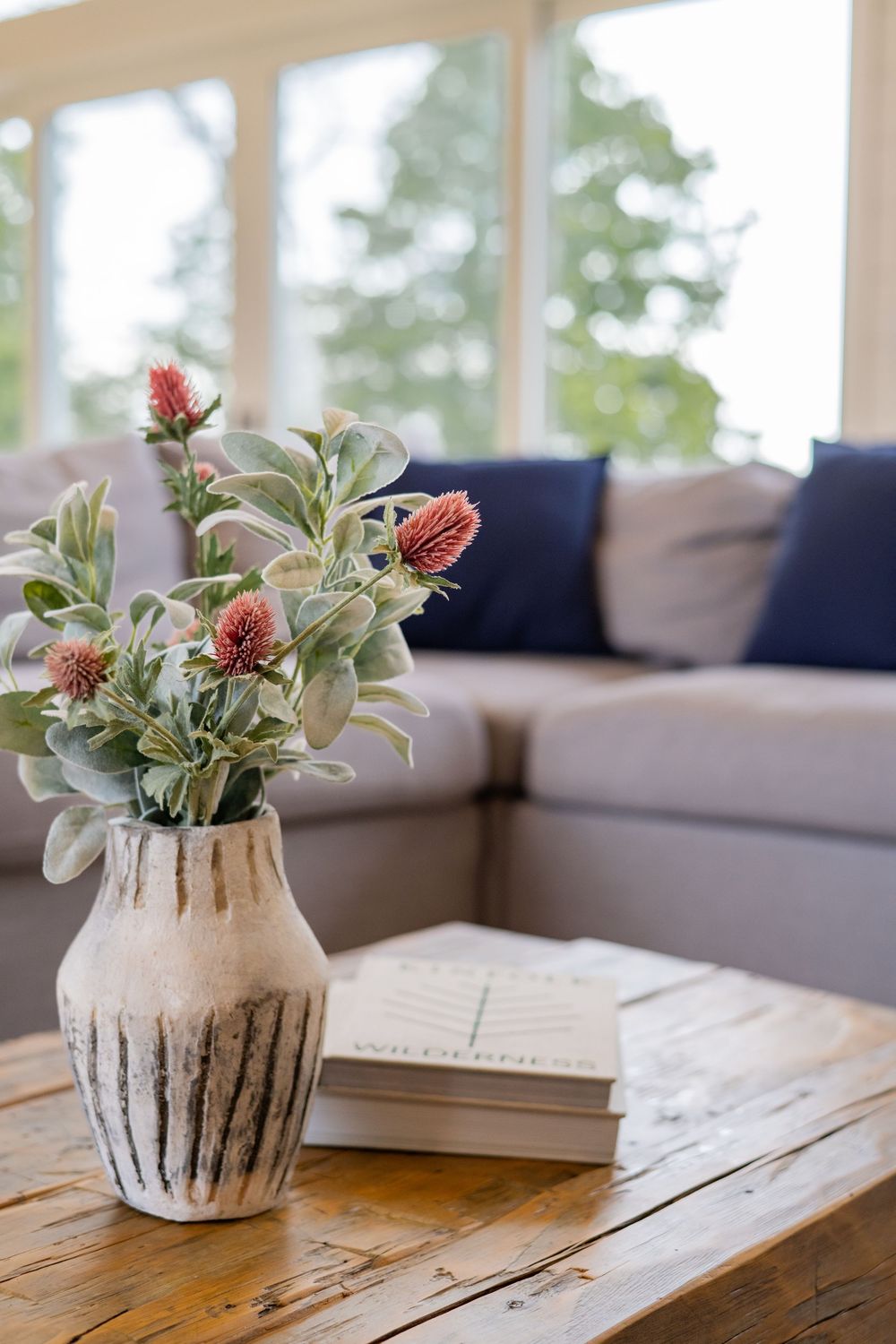 A rustic vase with pink flowers and greenery sits on a wooden table with books near a gray couch.