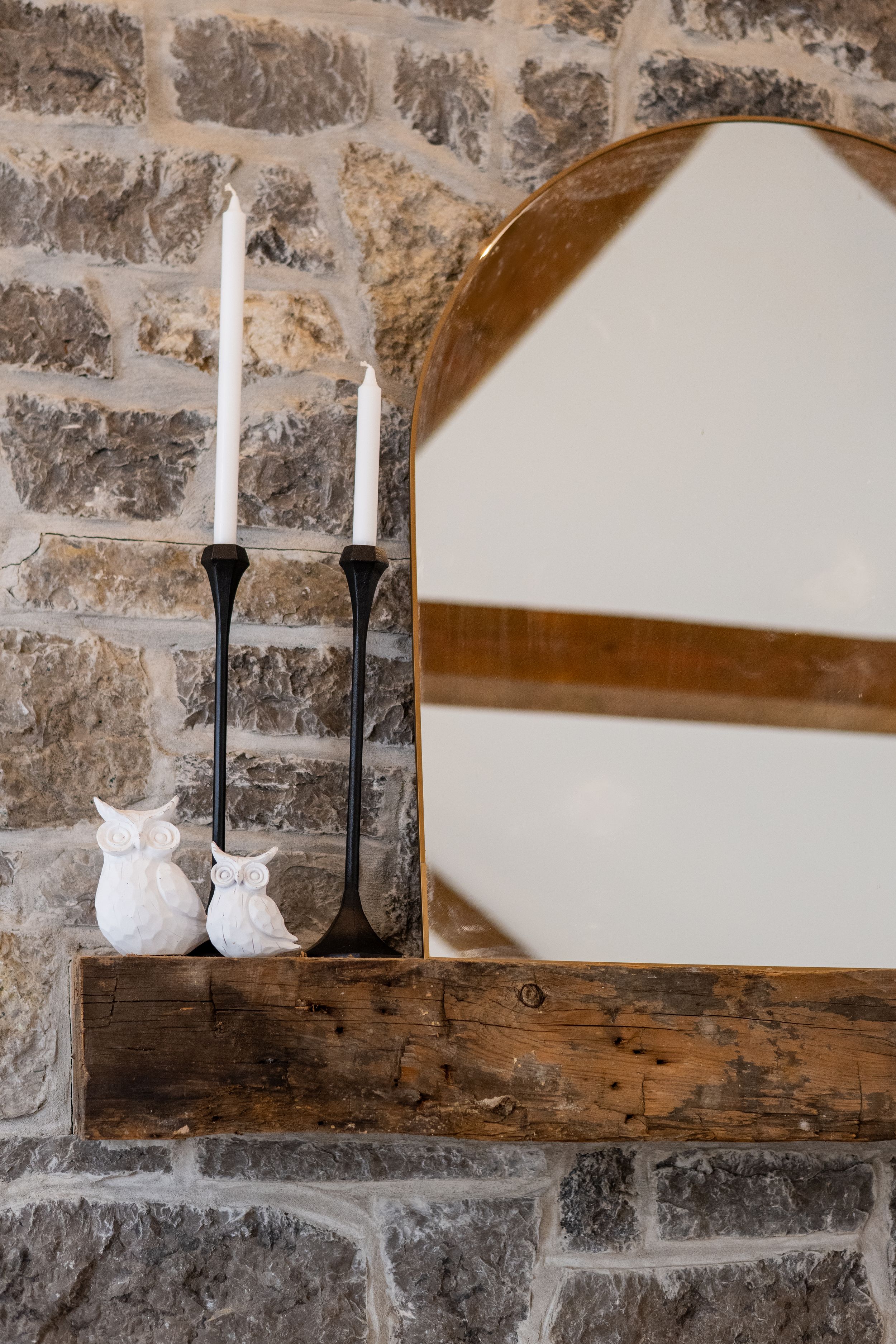 White ceramic owl figurines and black candlesticks on a wooden shelf against a stone wall.