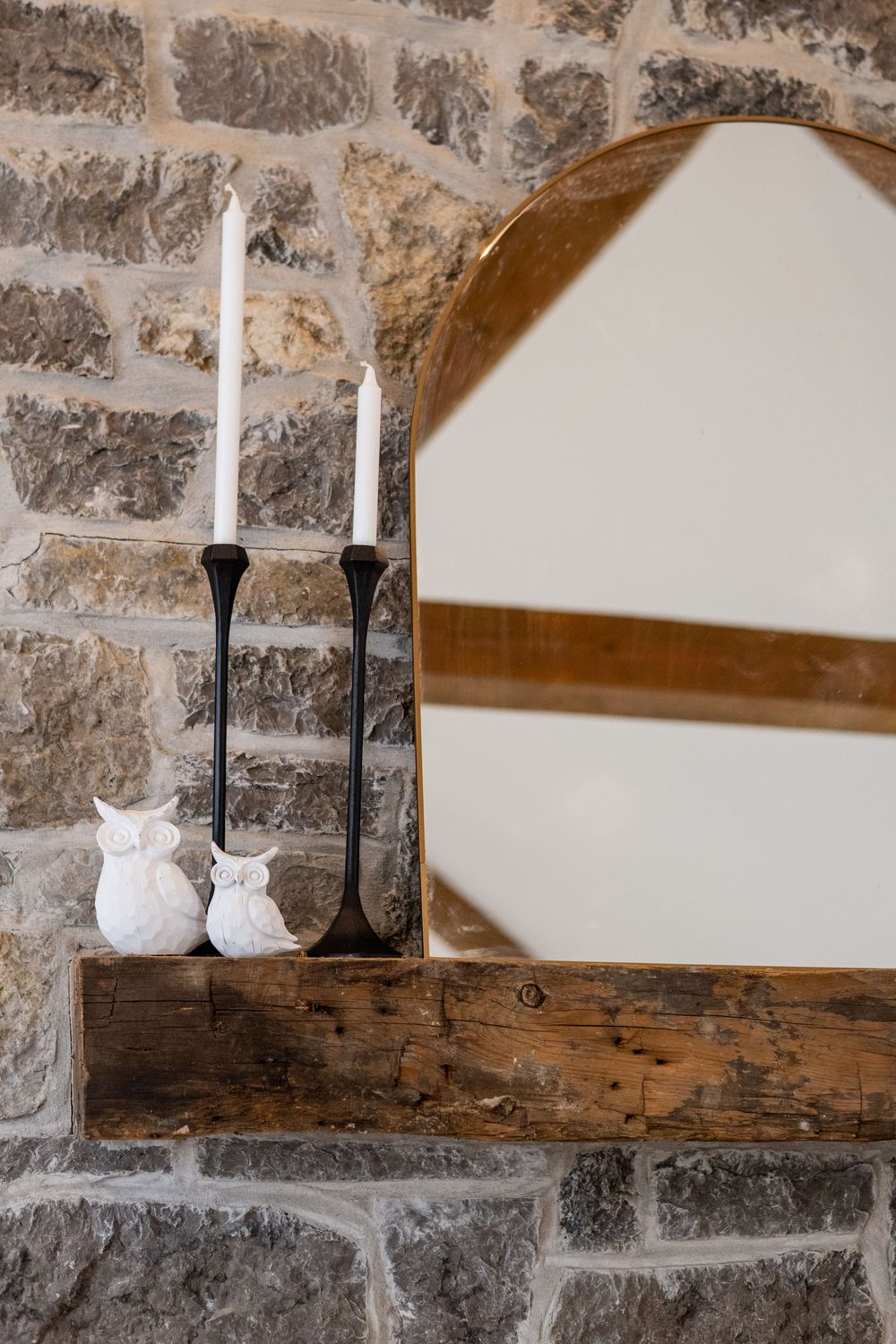 White ceramic owl figurines and black candlesticks on a wooden shelf against a stone wall.