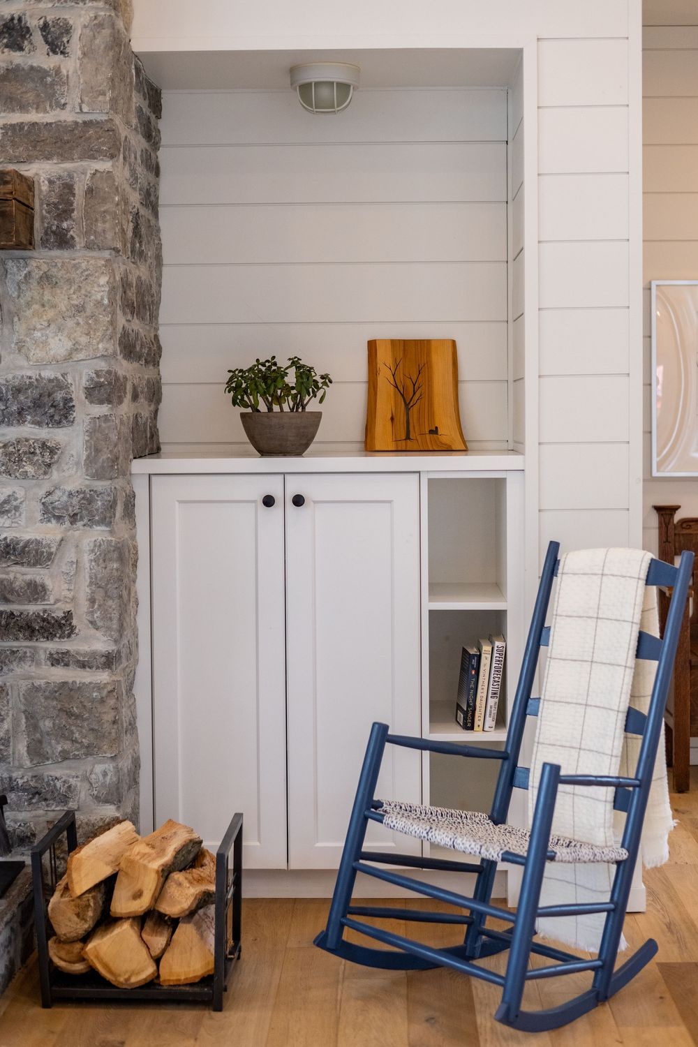 A blue rocking chair beside white cabinets and stone wall with firewood storage.