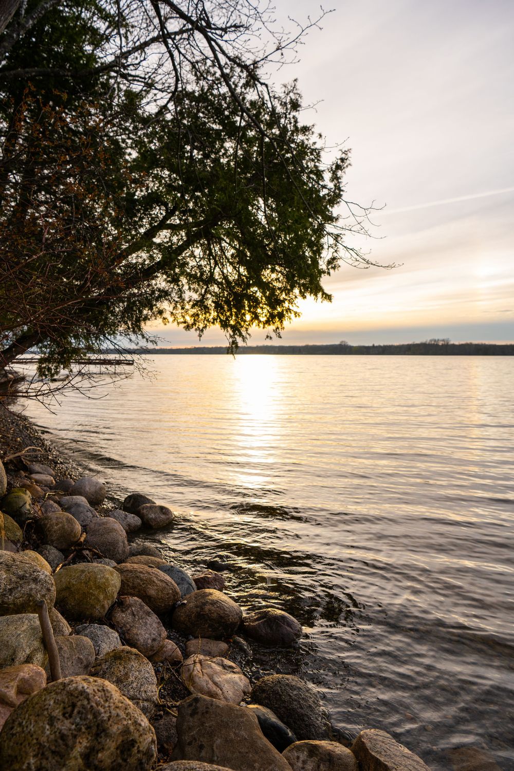 Sunset over a lake with rocks along the shoreline and overhanging tree branches.