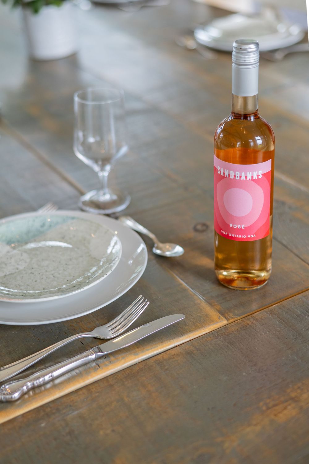 Table setting with white plates and pink wine bottle on rustic wooden surface.