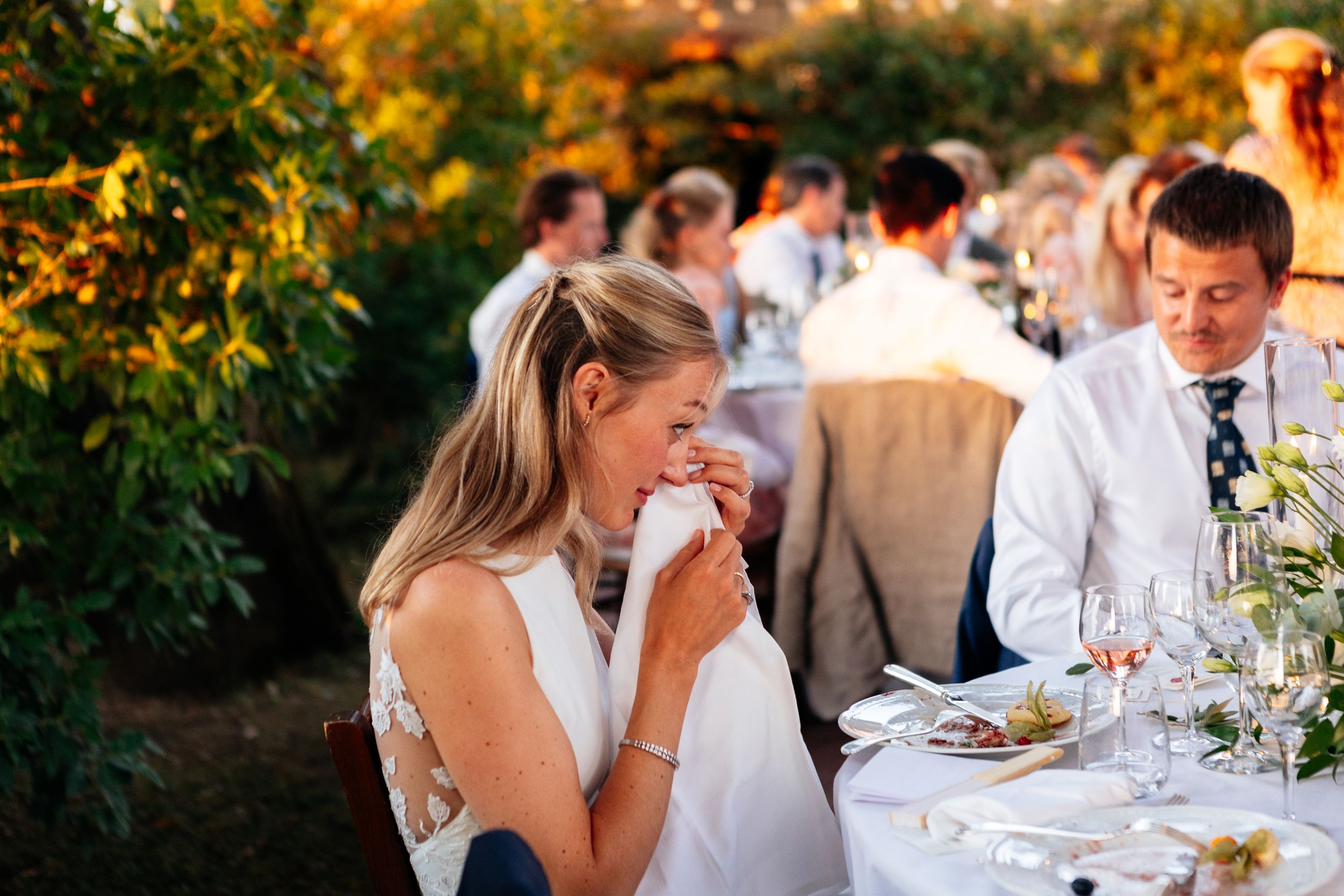 Guests enjoy an elegant outdoor dining experience at sunset in Tuscany while the bride wipes a tear away during the wedding speeches.