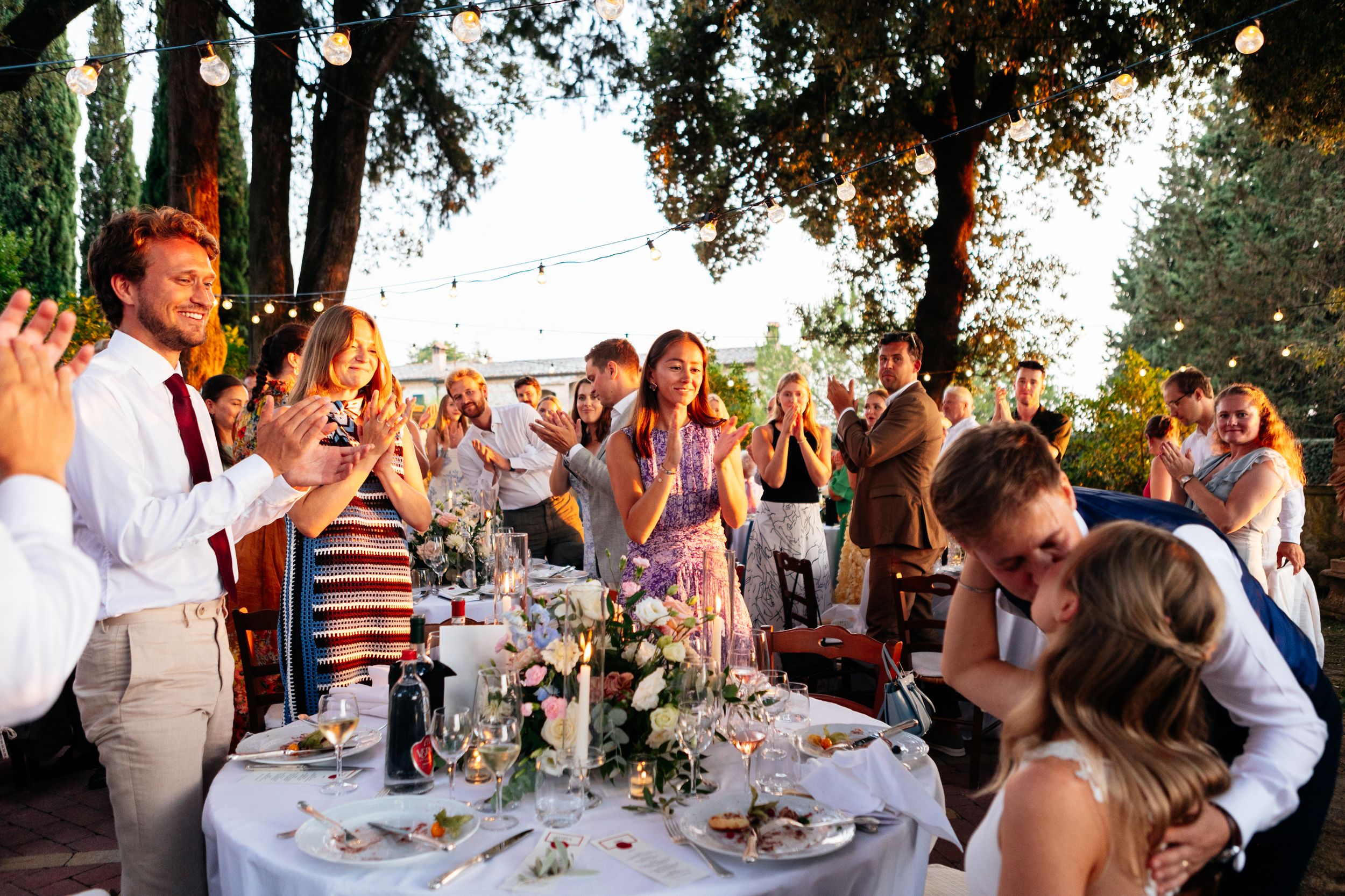 Wedding guests applaud during an outdoor reception with string lights as the bride and groom kiss.