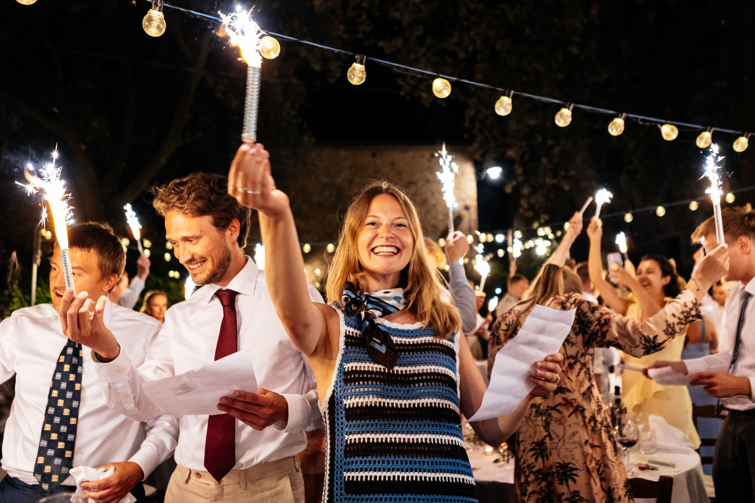 Wedding party guests celebrate with sparklers at a festive outdoor evening reception under string lights in Tuscany.