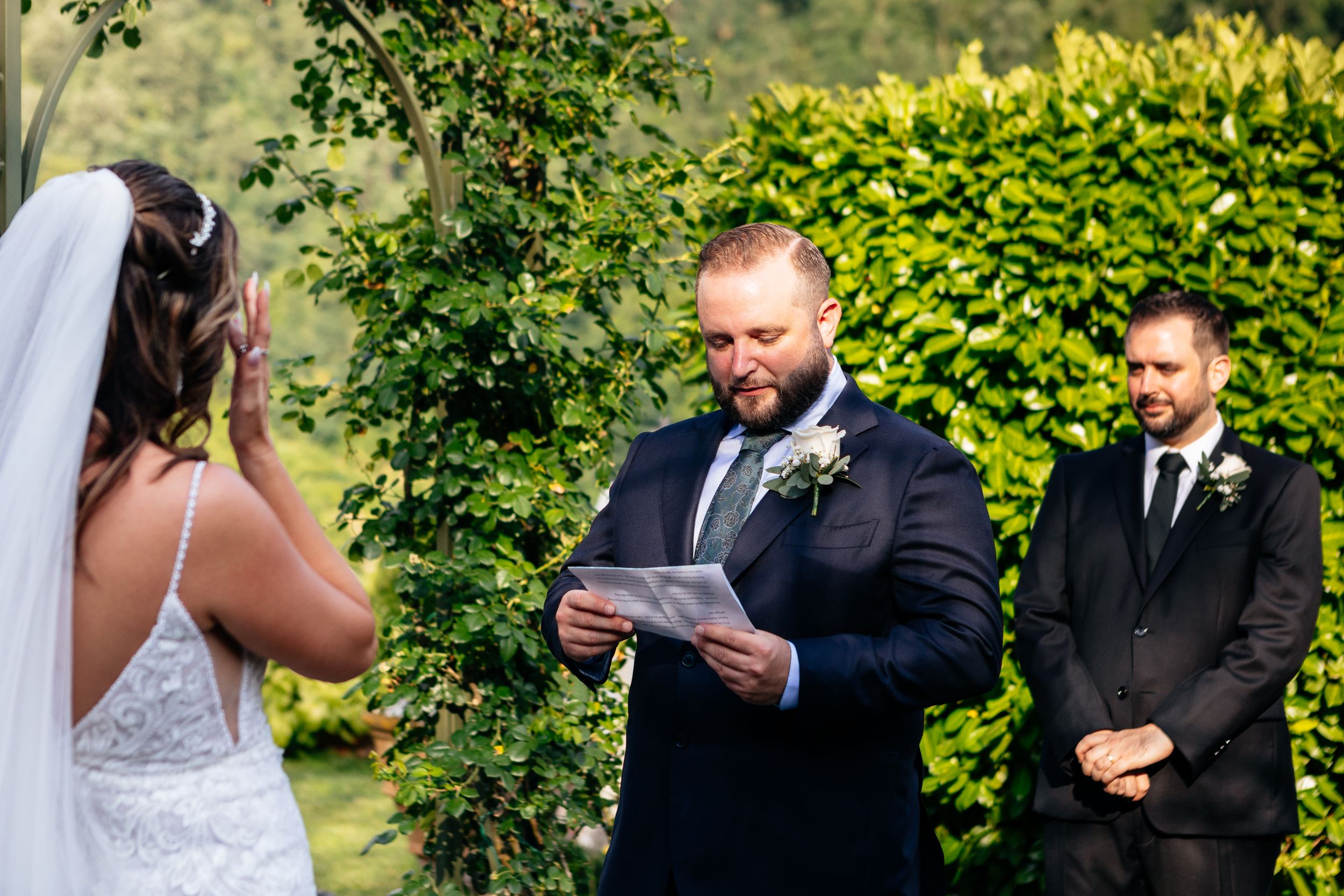 A wedding ceremony takes place outdoors with participants standing in front of lush green hedges in a garden setting at Burlamacchi Villas