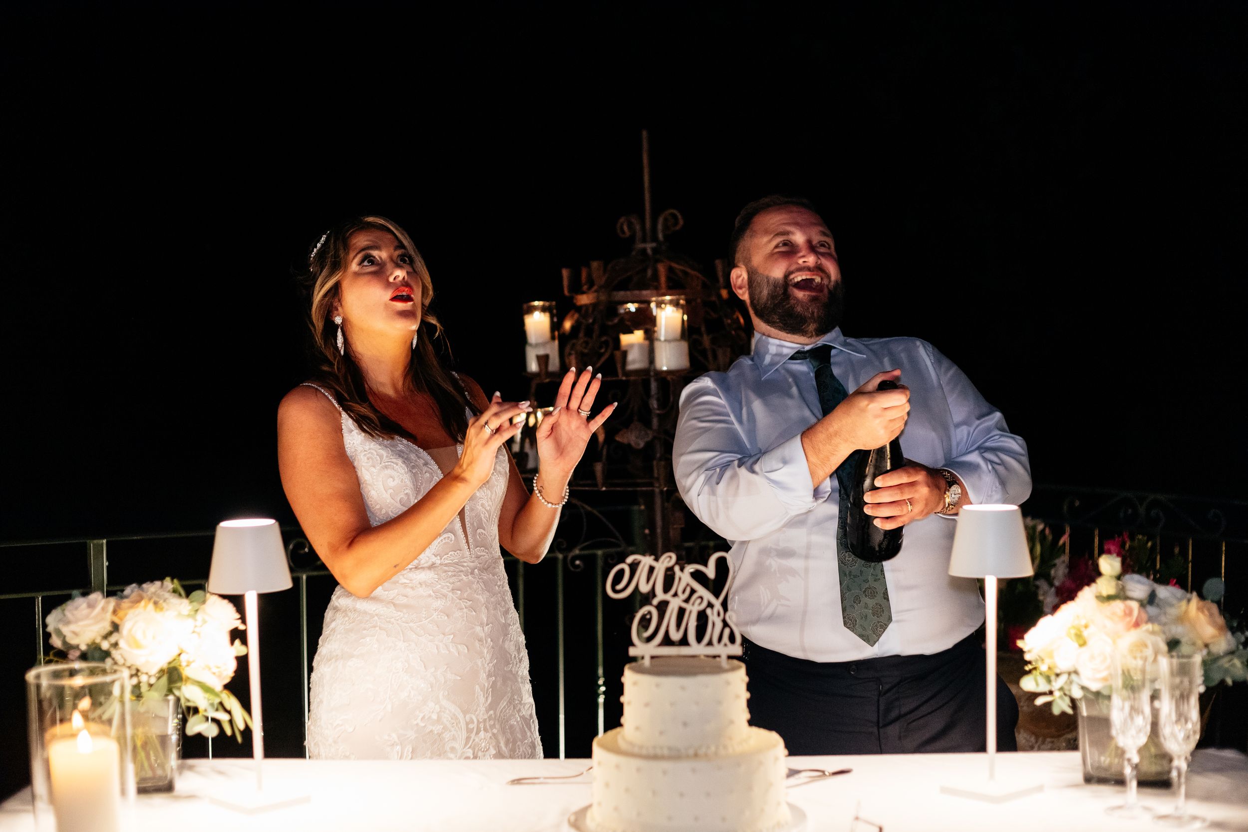Two people in wedding attire laugh while raising champagne glasses during a toast at their reception beside a white cake.
