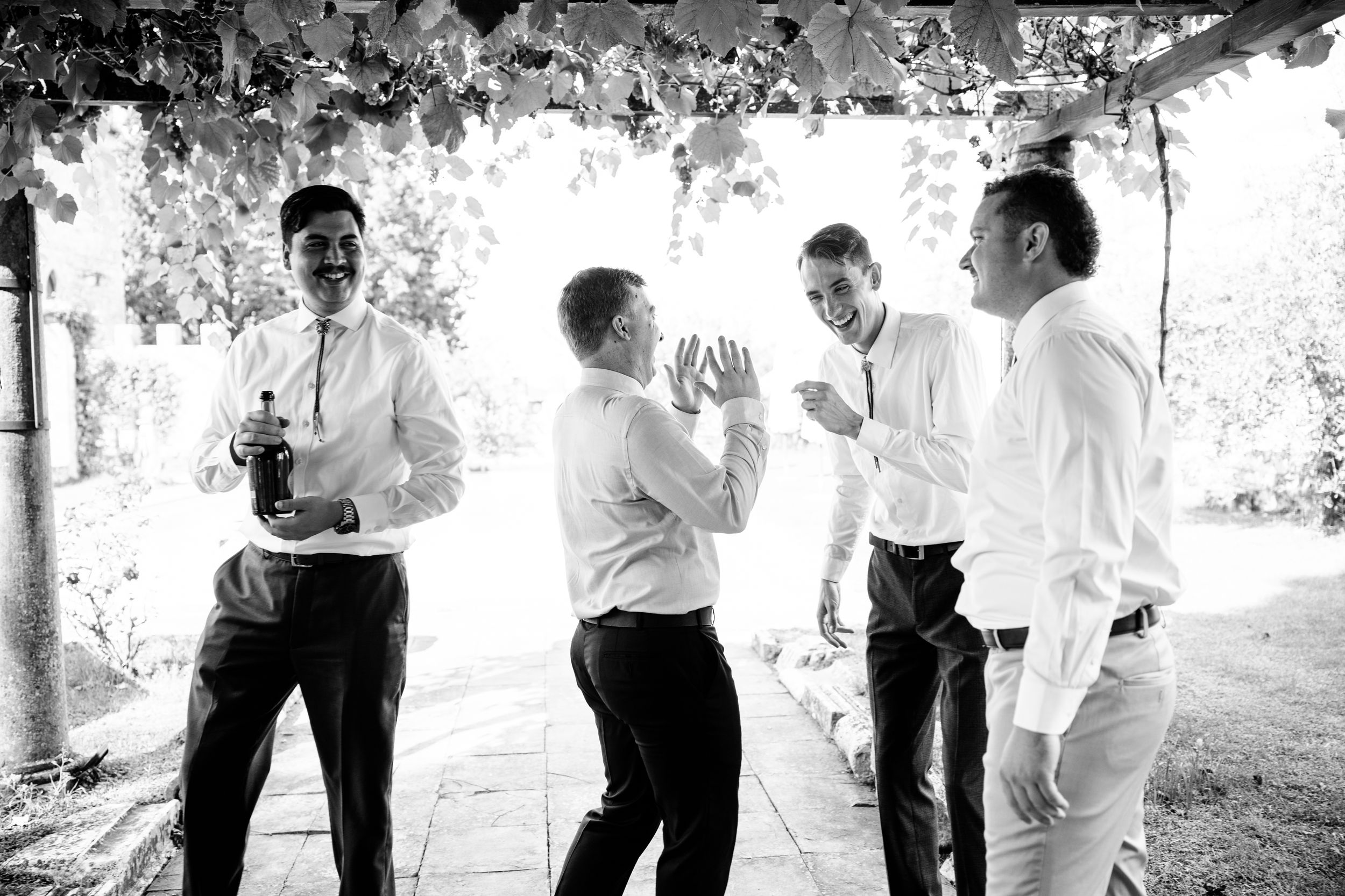 Groomsmen in formal white attire laughing together under an outdoor canopy in black and white photography in Tuscany.