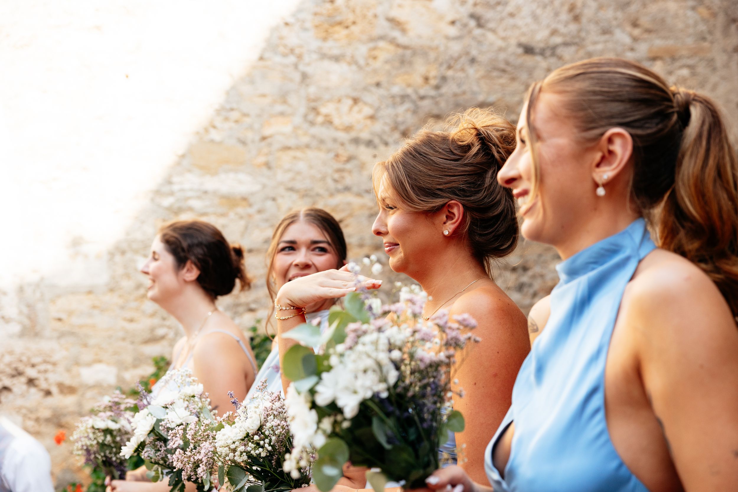 Bridesmaids in formal attire stand in a row holding white floral bouquets while smiling outdoors near a stone wall.