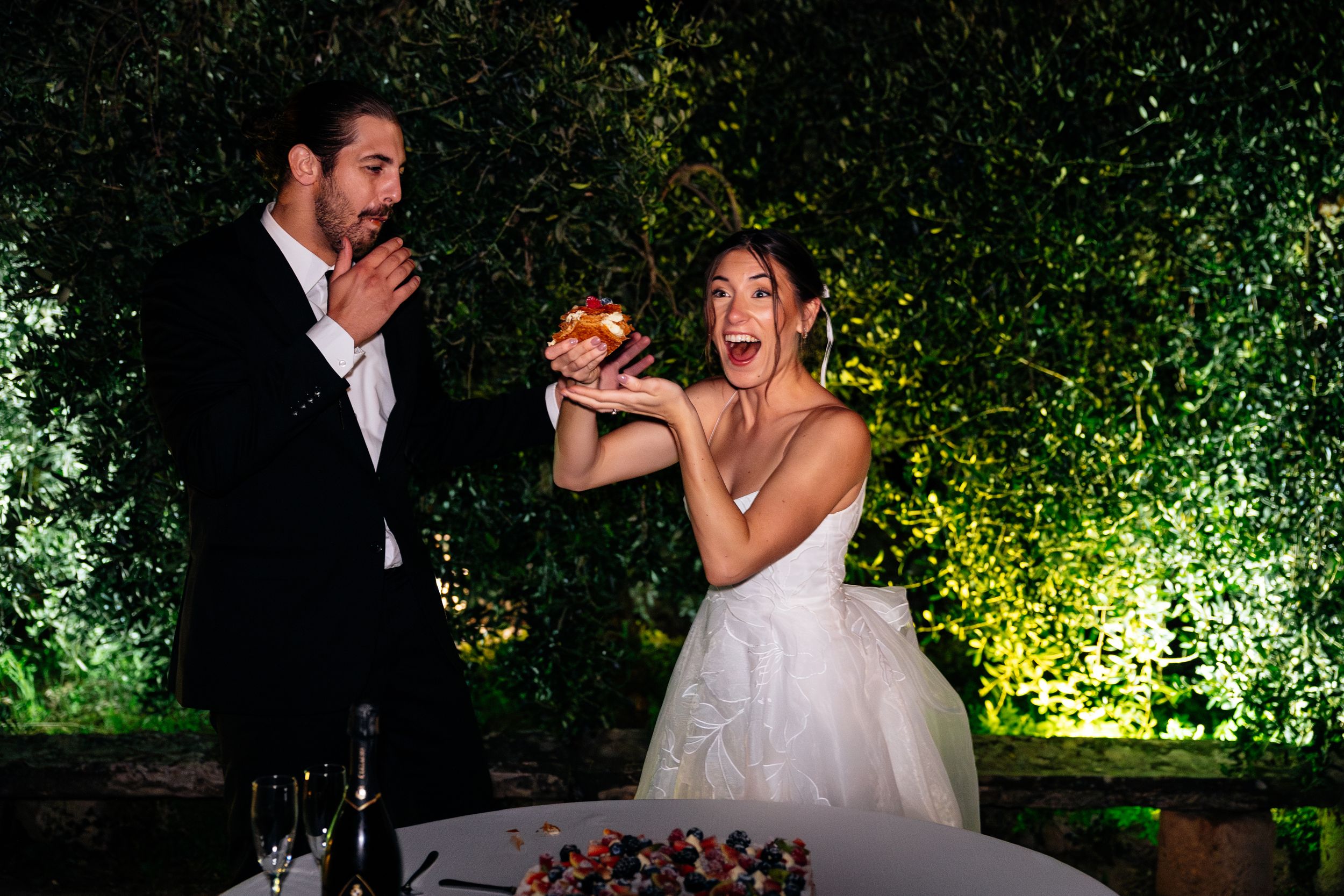 A couple reacts playfully while cutting their wedding cake together at an outdoor reception with illuminated greenery in Tuscany.