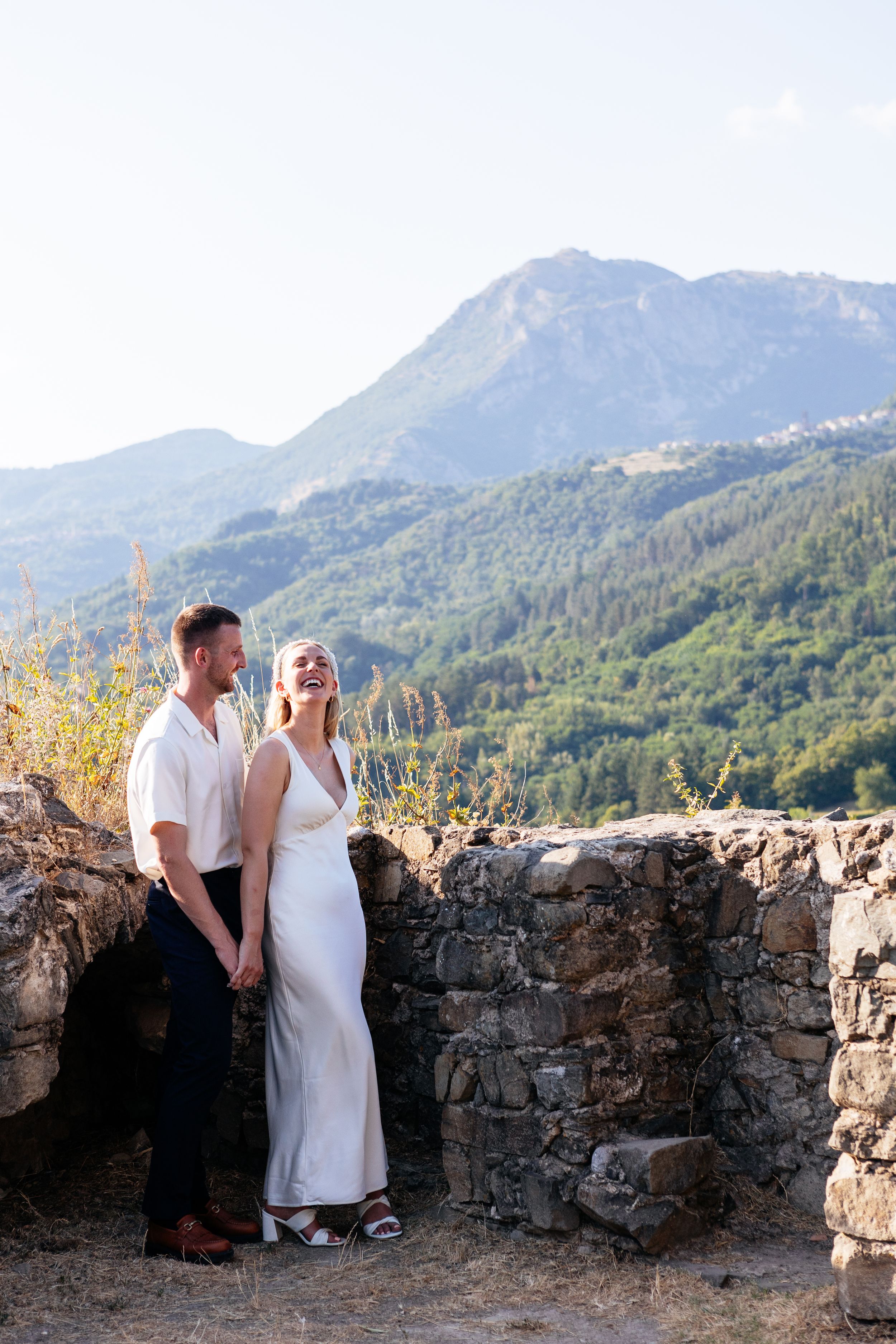 Two people in elegant wedding outfits enjoy a romantic moment against a backdrop of majestic mountains and stone walls in the Garfagnana.