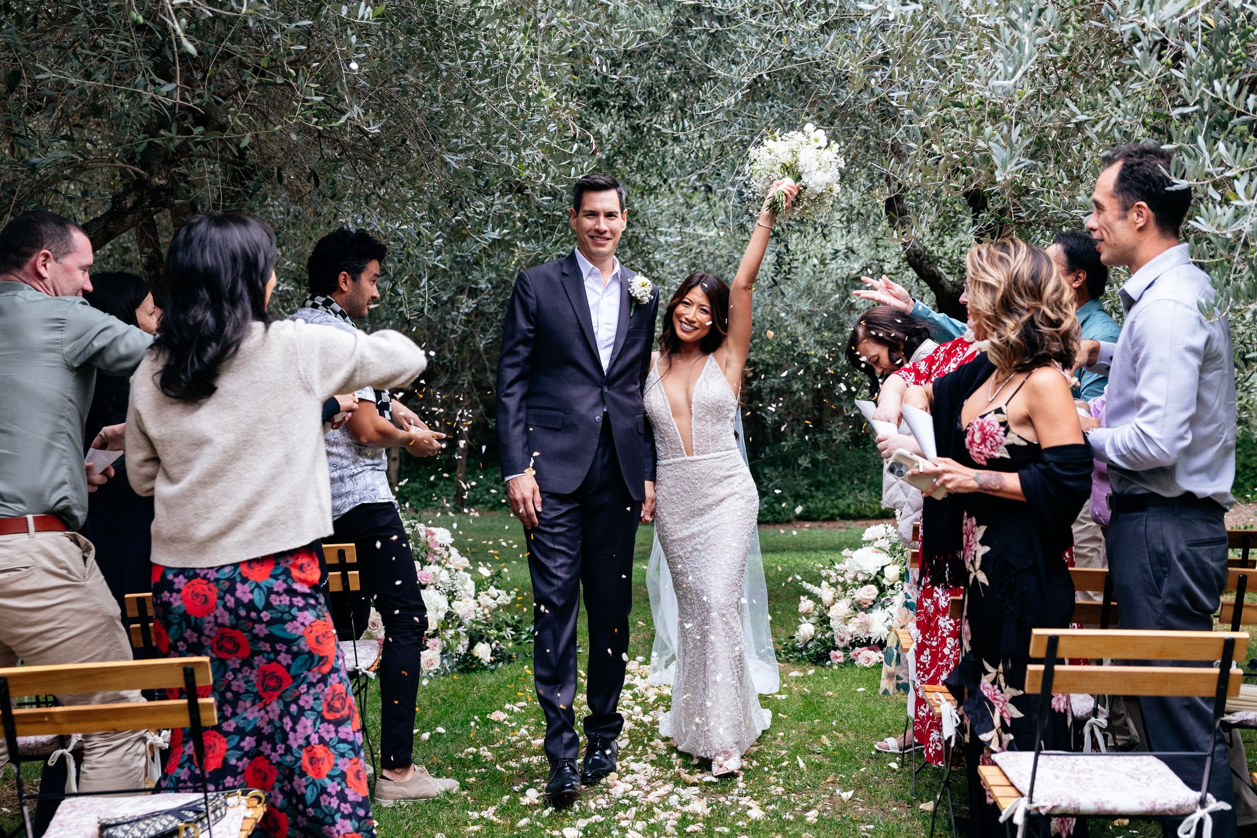 Wedding guests toss white flower petals as newlyweds walk down the aisle during an outdoor garden ceremony in Florence.