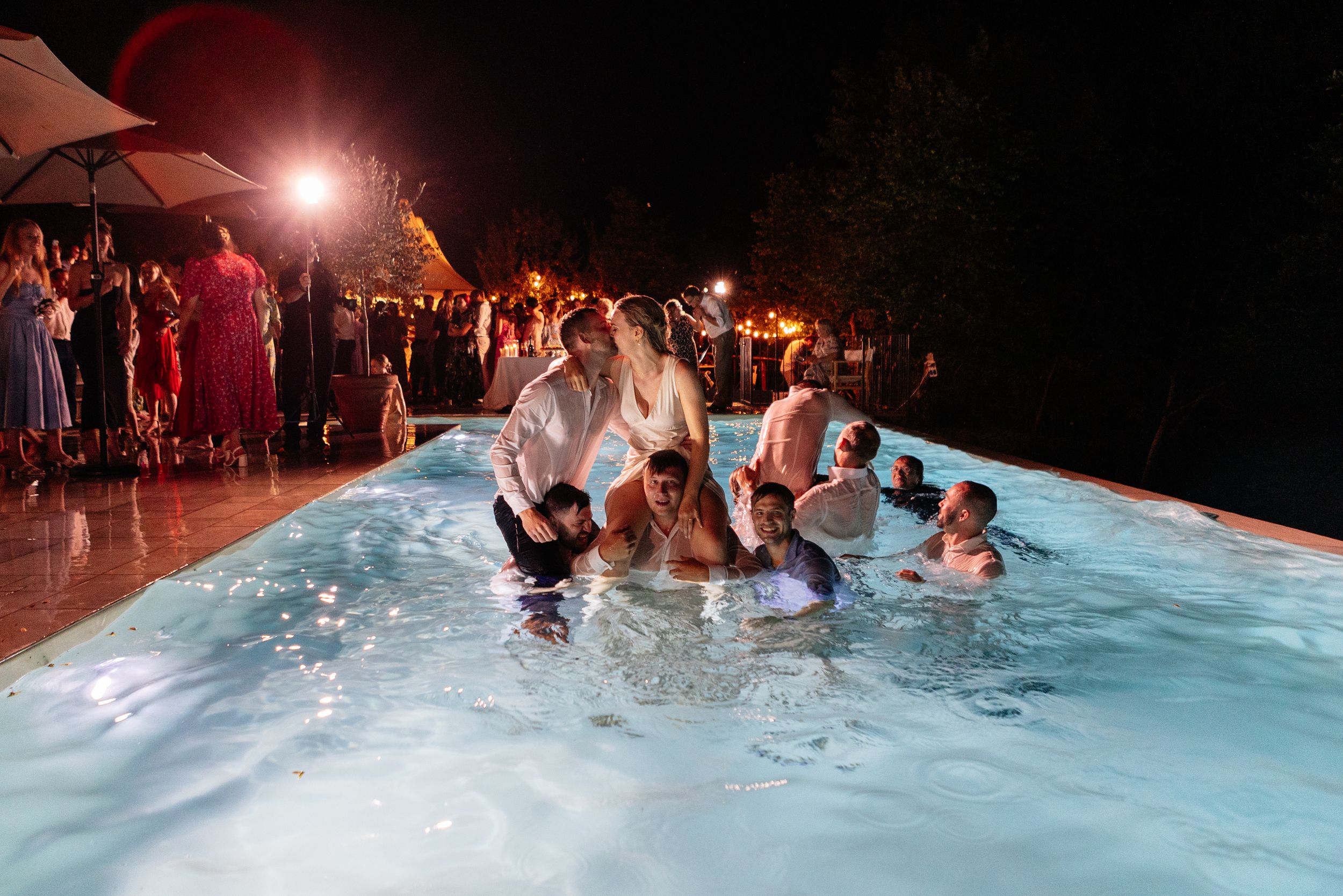 Wedding party guests splash and play in an illuminated pool at night while others watch from under umbrellas on the deck.