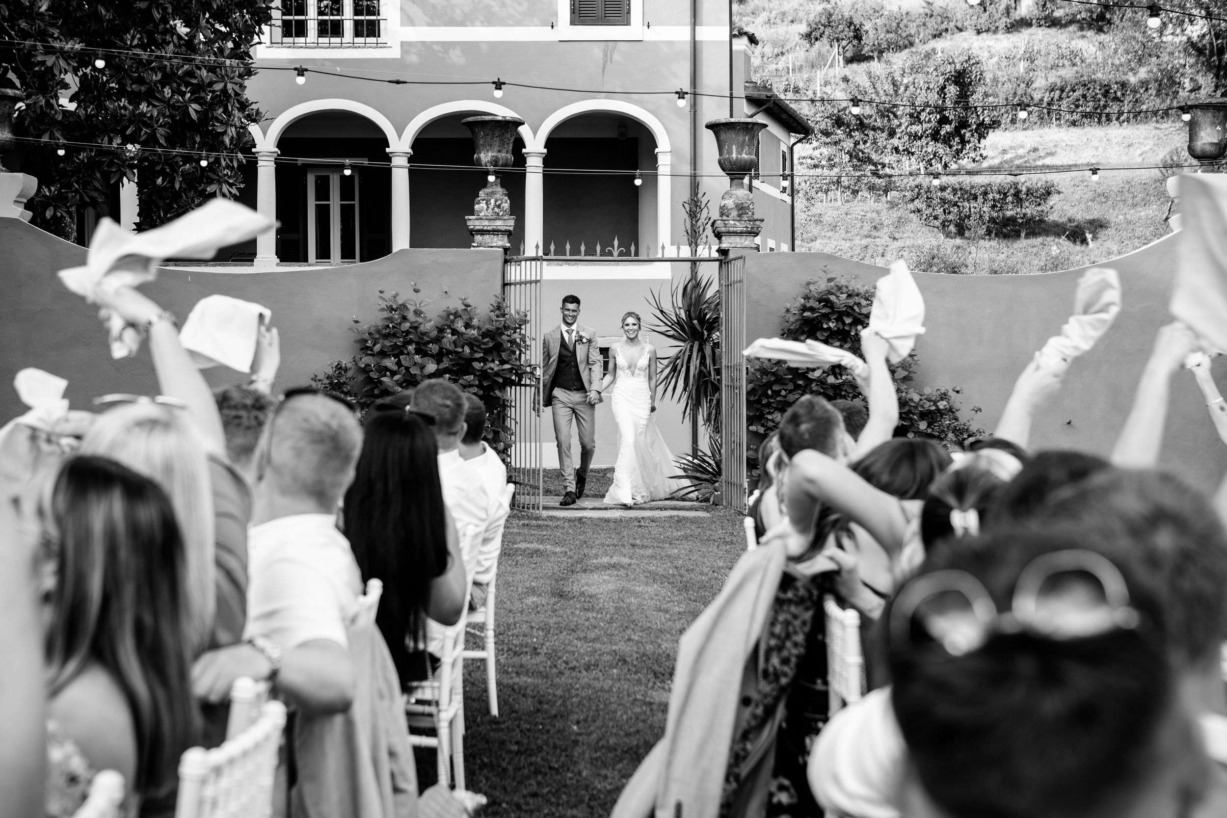 Guest wave their napkins in the air as the newly married couple walk in to the outdoor dinner of their destination wedding in Barga Tuscany