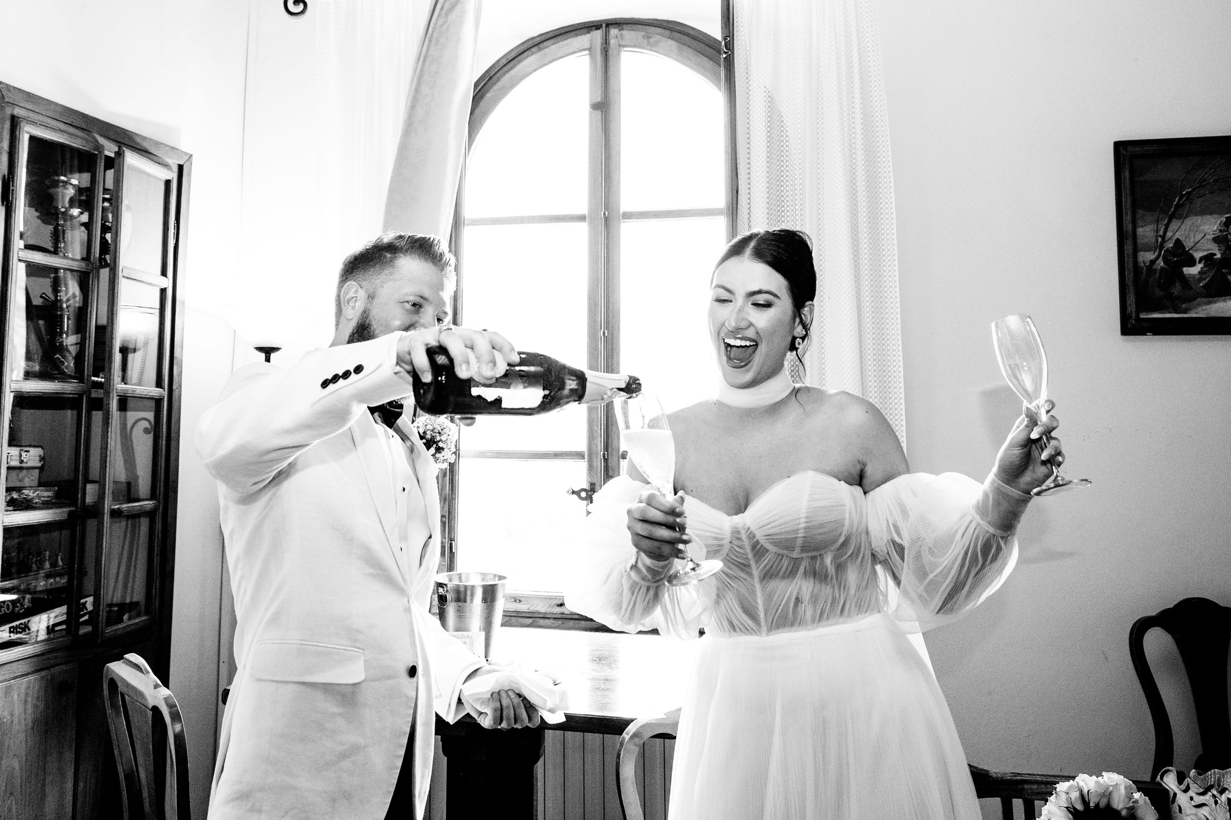 A joyful celebration after getting married as Prosecco is being poured in an elegant room with an arched window in Tuscany.