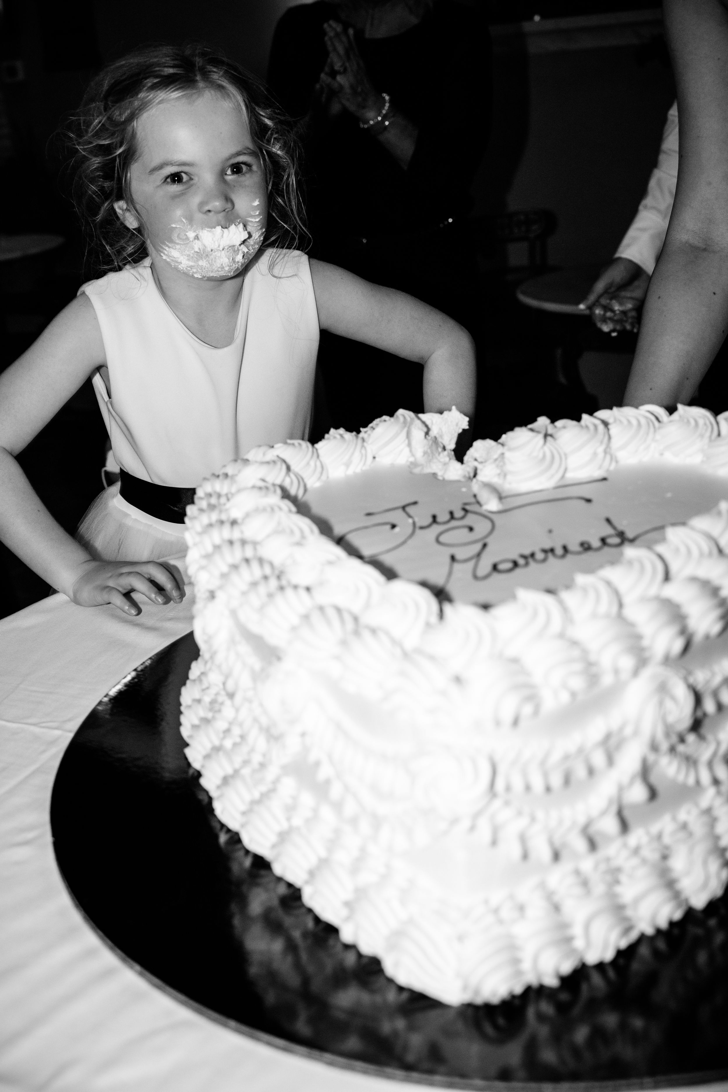 A black and white photo of a flower girl with cake with white frosting and whipped cream on her face after taking a bit out of it.
