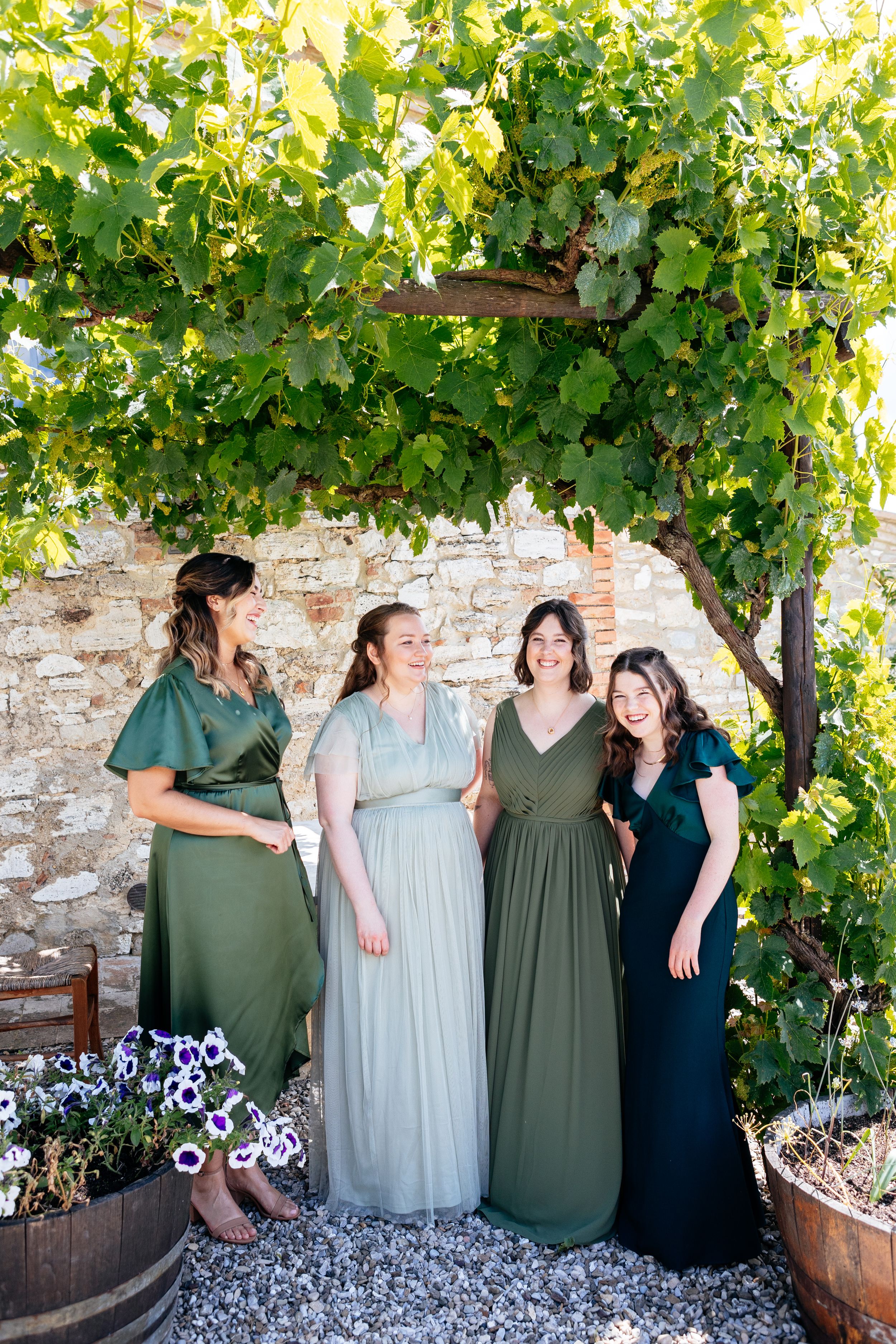 Bridesmaids in different but coordinating long dresses stand together under grapevines at a rustic stone wall with flower planters.