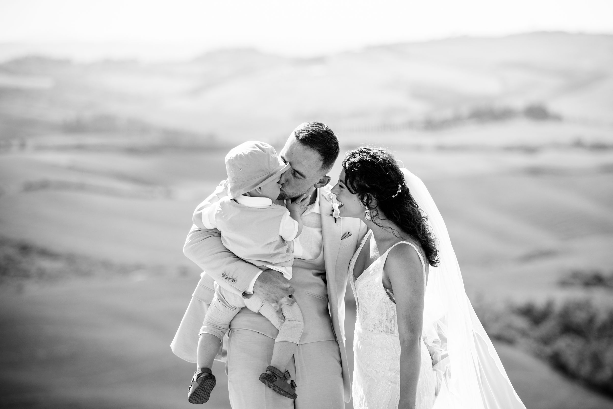 A couple in wedding attire share a tender moment with their child against a scenic hillside backdrop in Tuscany in black and white.