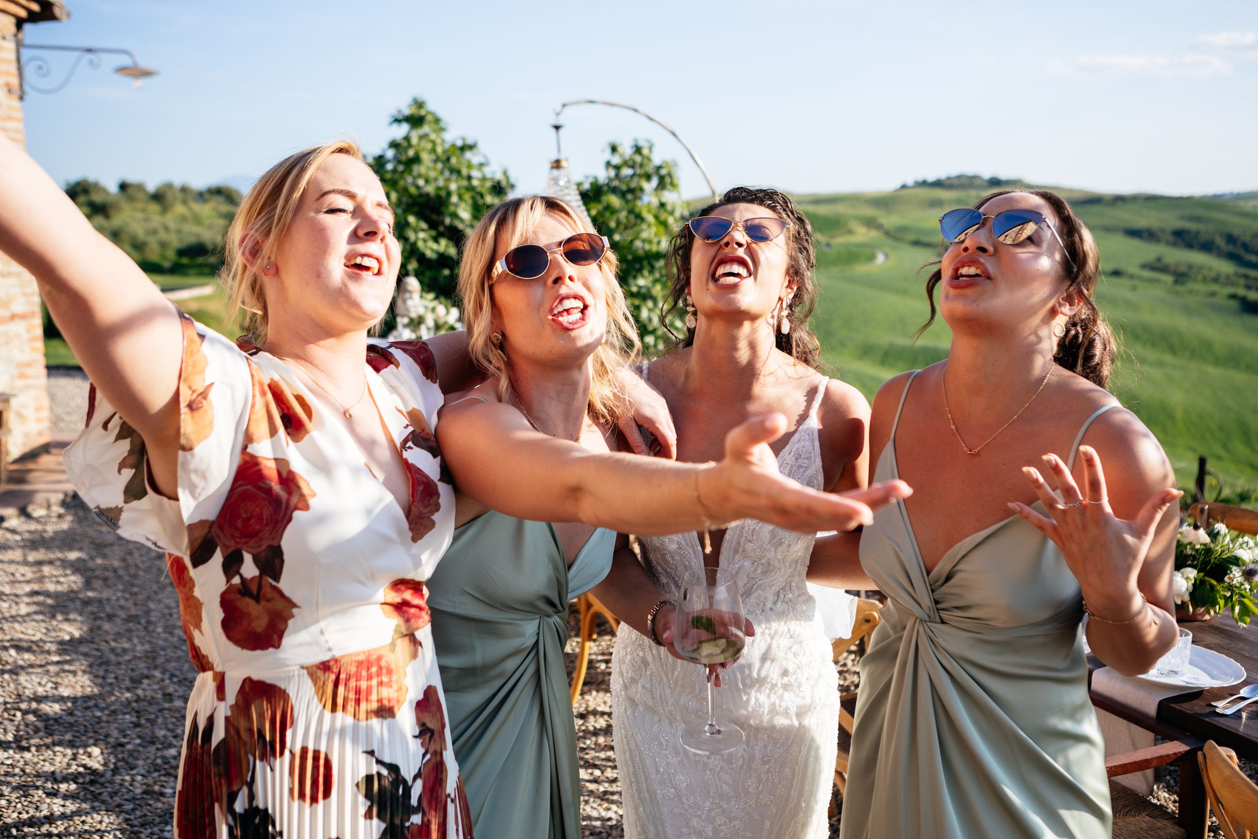 A group celebrates joyfully at wedding against a scenic green hillside backdrop on a sunny summer day in Tuscany.