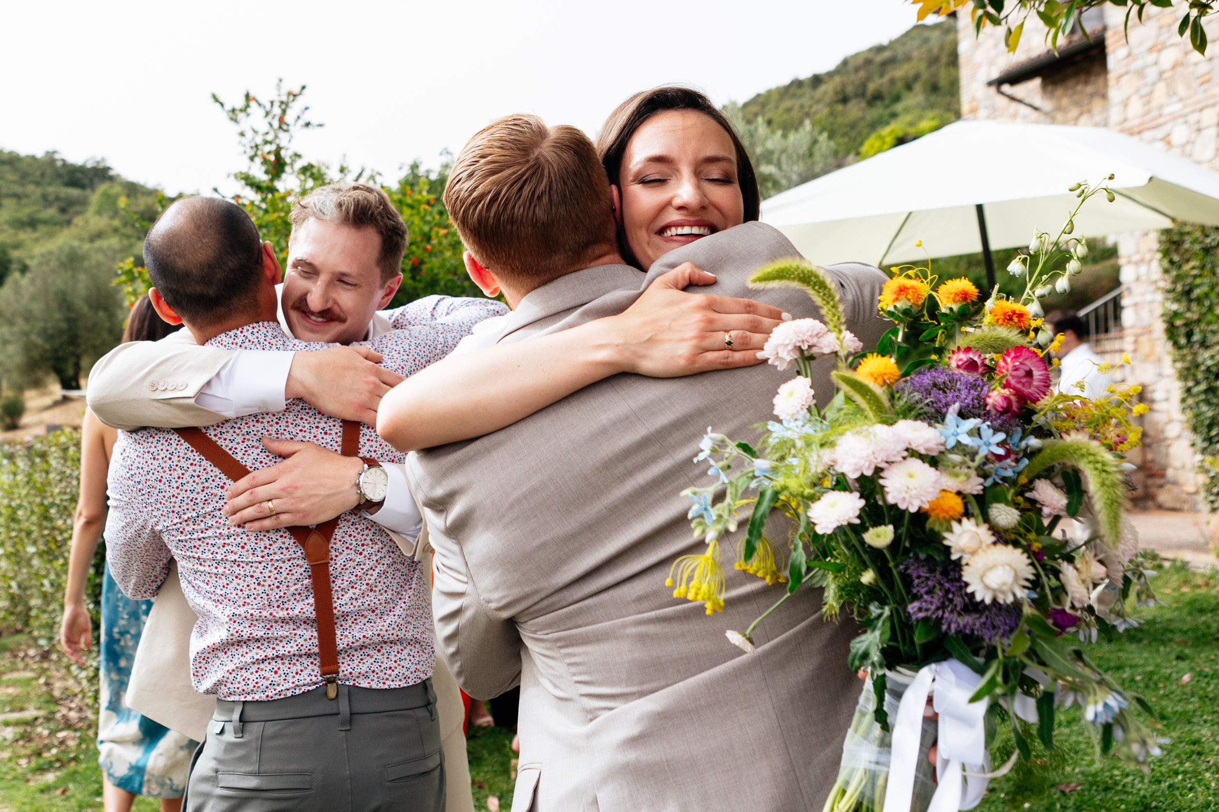 Joyful group embraces at an outdoor wedding reception with colorful floral arrangements and white umbrellas in background in Umbria.