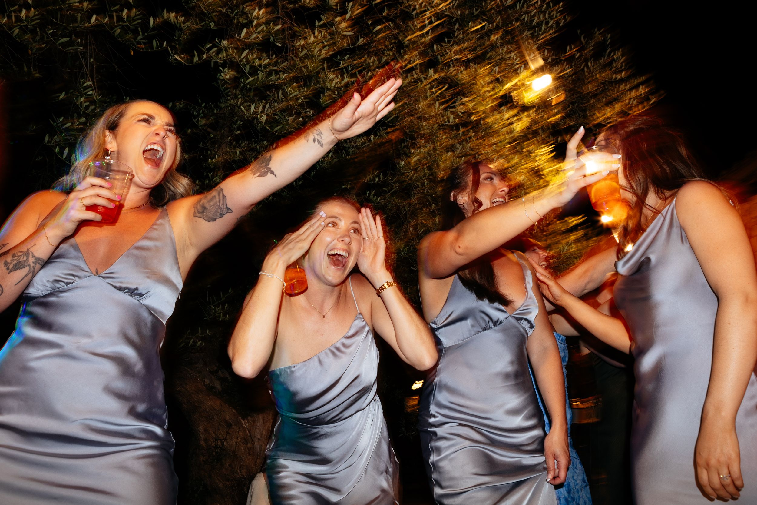 Bridesmaids in matching blue satin dresses react with dramatic expressions during a nighttime celebration.