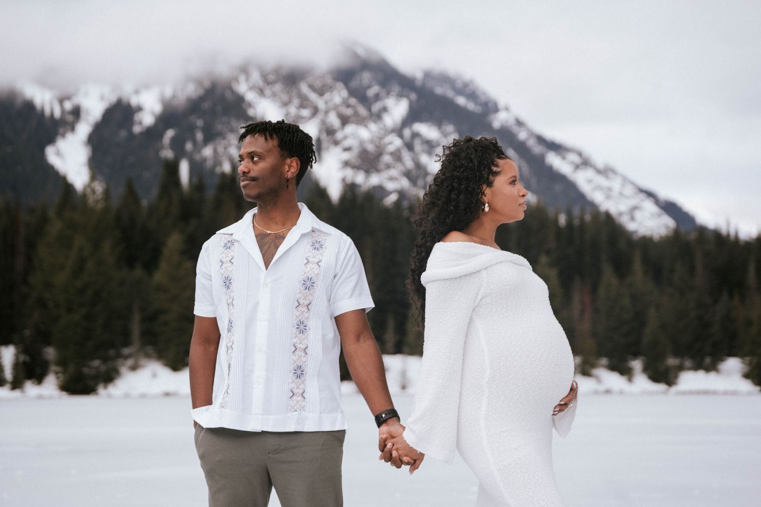 Pregnant couple in white holding hands with snowy mountains and pine trees behind them.