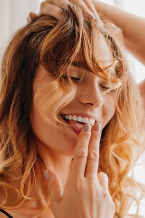 A person with wavy blonde hair smiles and touches their lips with their fingers in warm lighting.