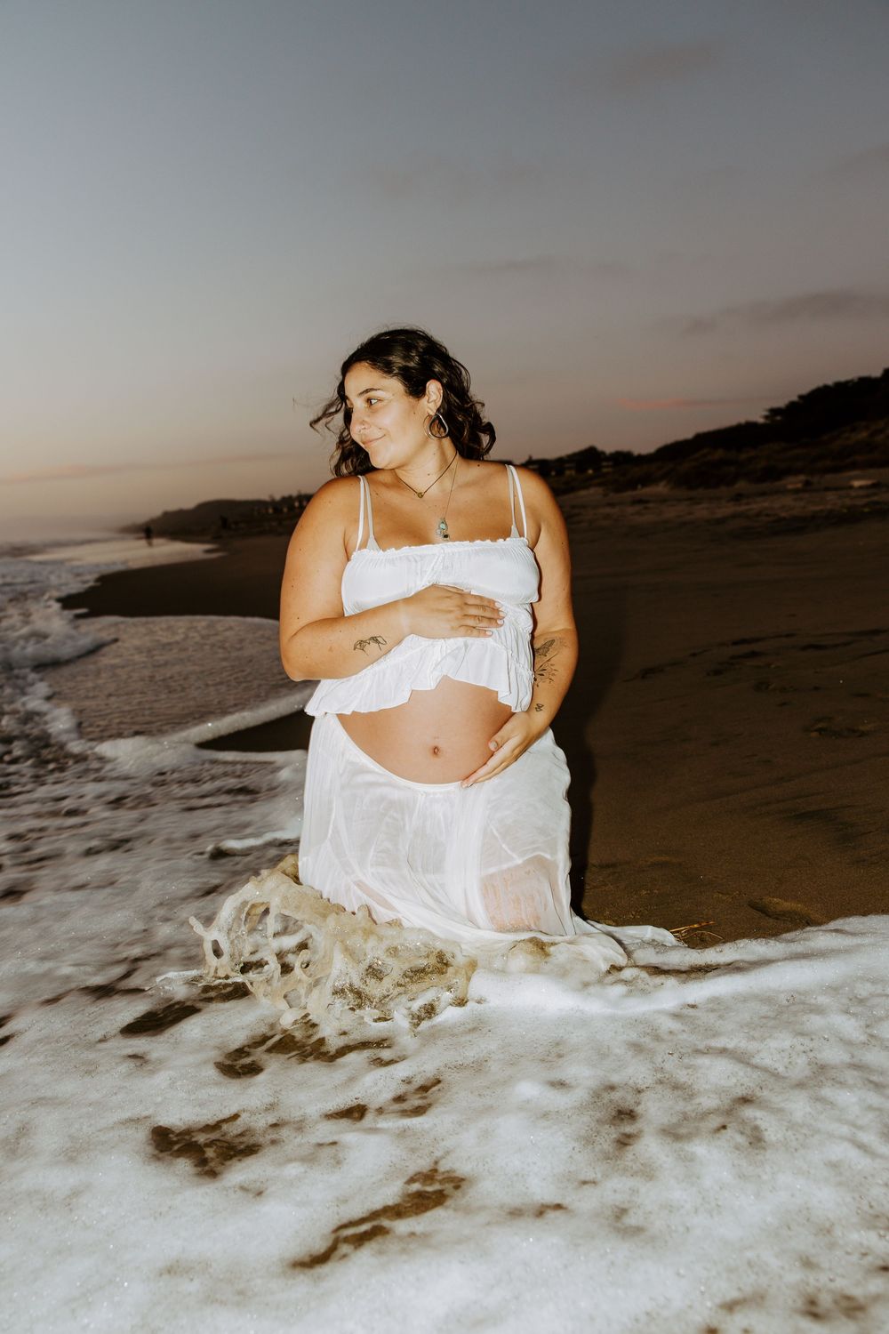 A person in white clothing poses on a beach at sunset as waves crash against the shore.