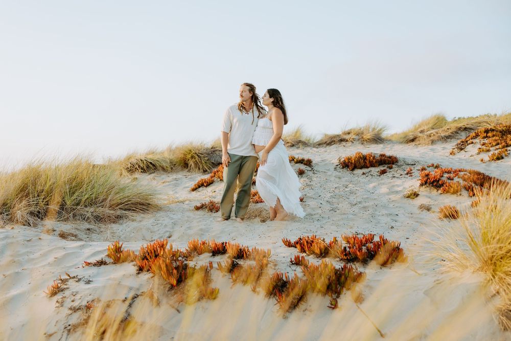 A couple sits together on sandy beach dunes at sunset while enjoying a romantic coastal view.