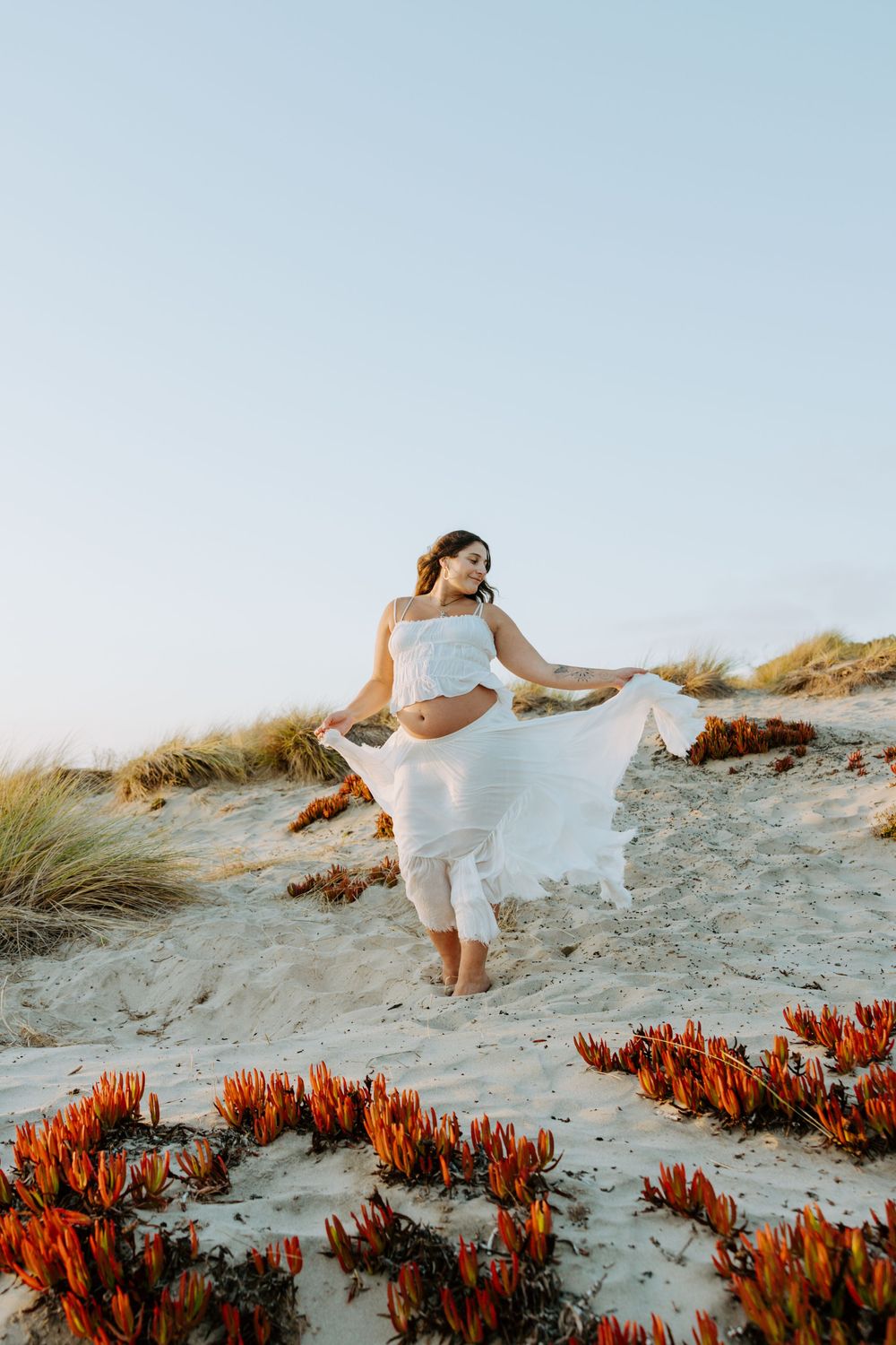 A person in a flowing white dress stands gracefully among orange succulents on a sandy beach at sunset.