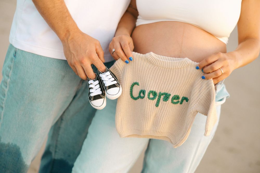 A person holds small baby shoes and a knit sweater against a light blue background.