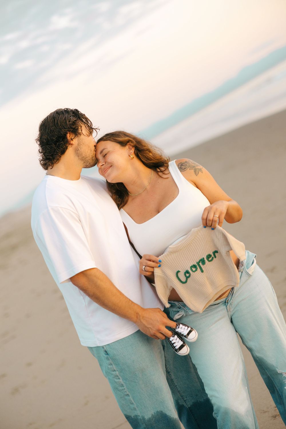 A couple in casual white and beige outfits embraces romantically on a misty beach during a photoshoot.