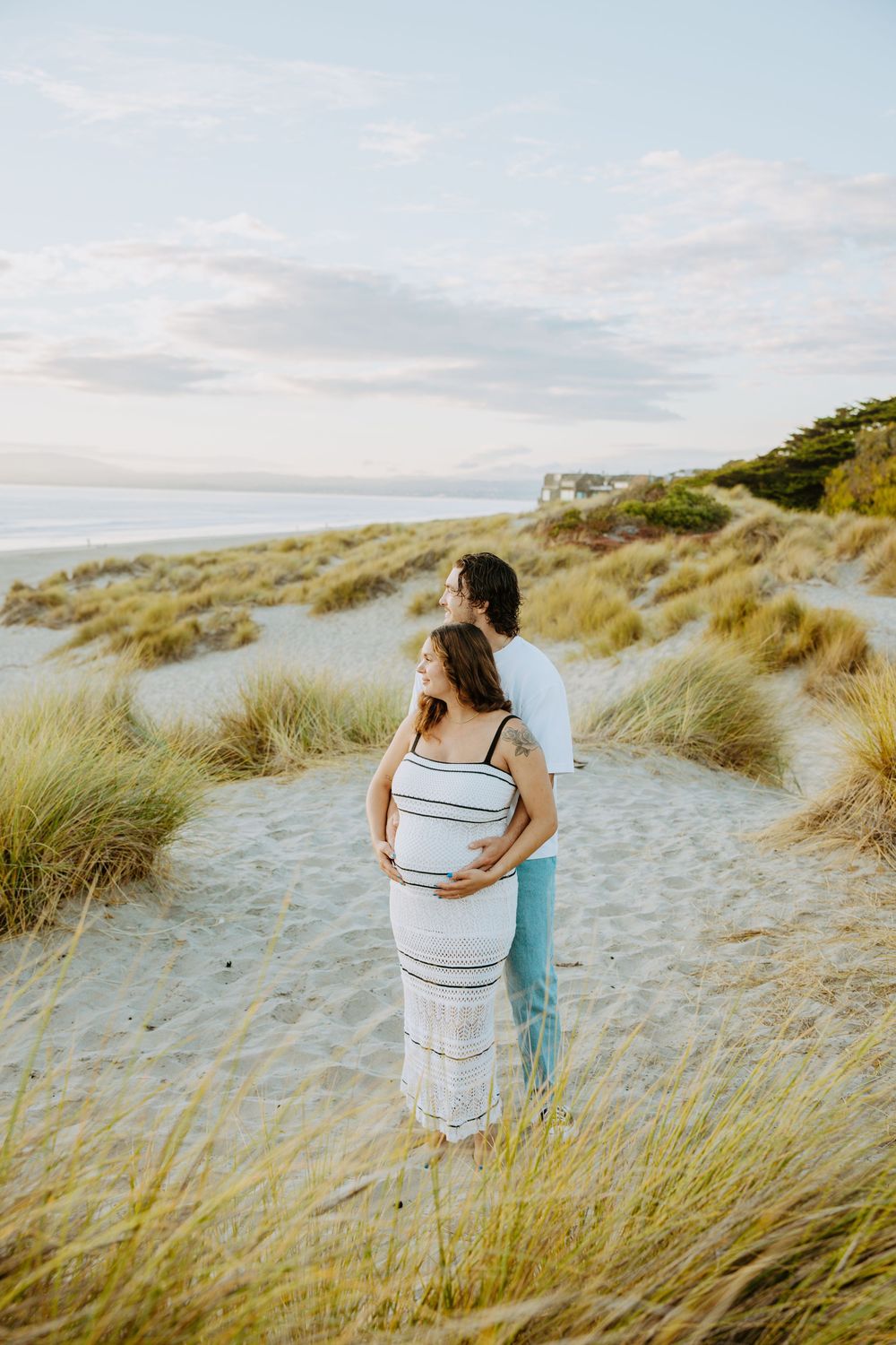 A couple embraces on a sandy coastal path surrounded by tall beach grass during golden hour.