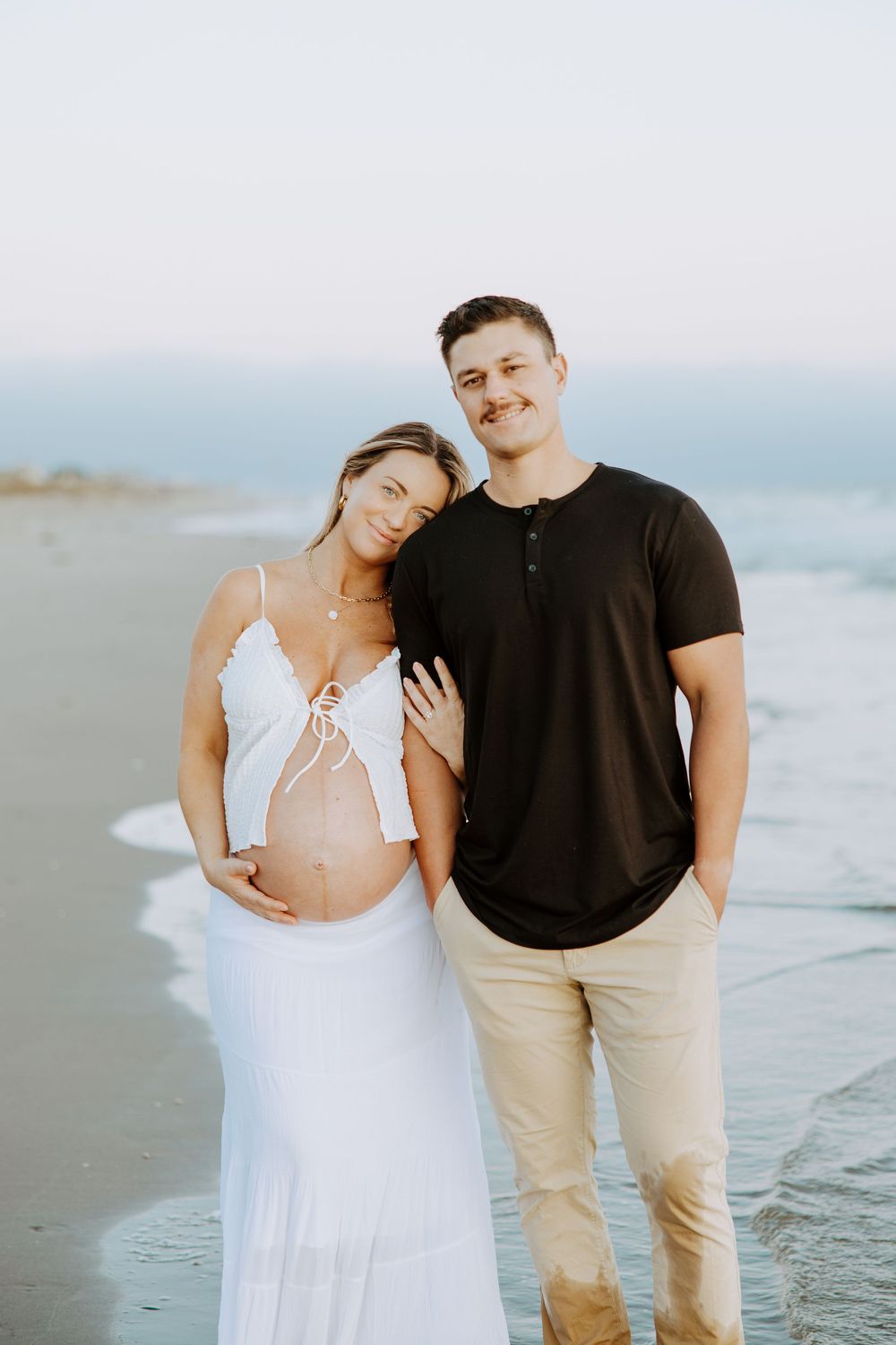 Couple in white dress and black shirt poses lovingly together on a serene beach at sunset.