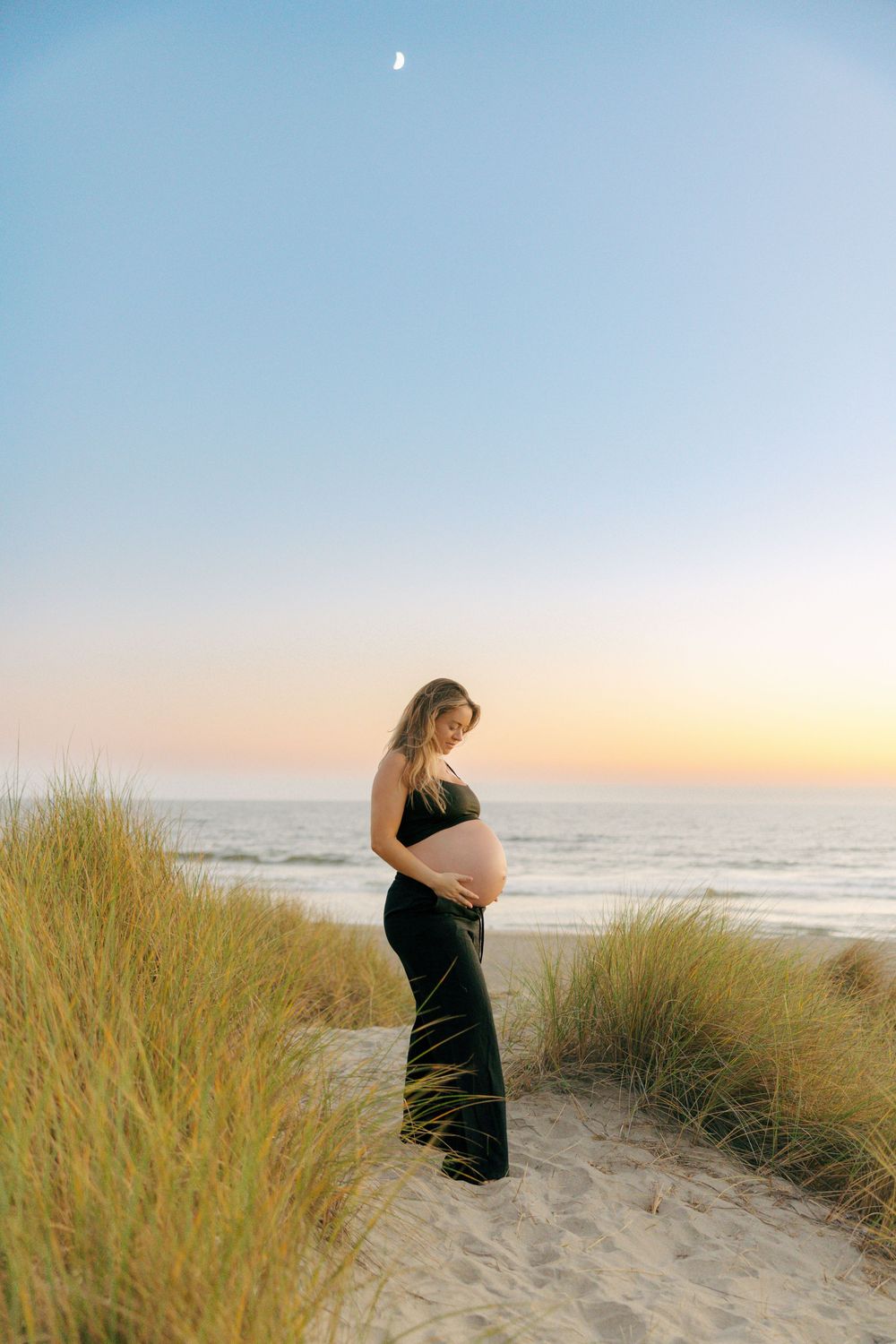 A silhouetted pregnant figure stands on sandy dunes at sunset with beach grass and a crescent moon in the sky.