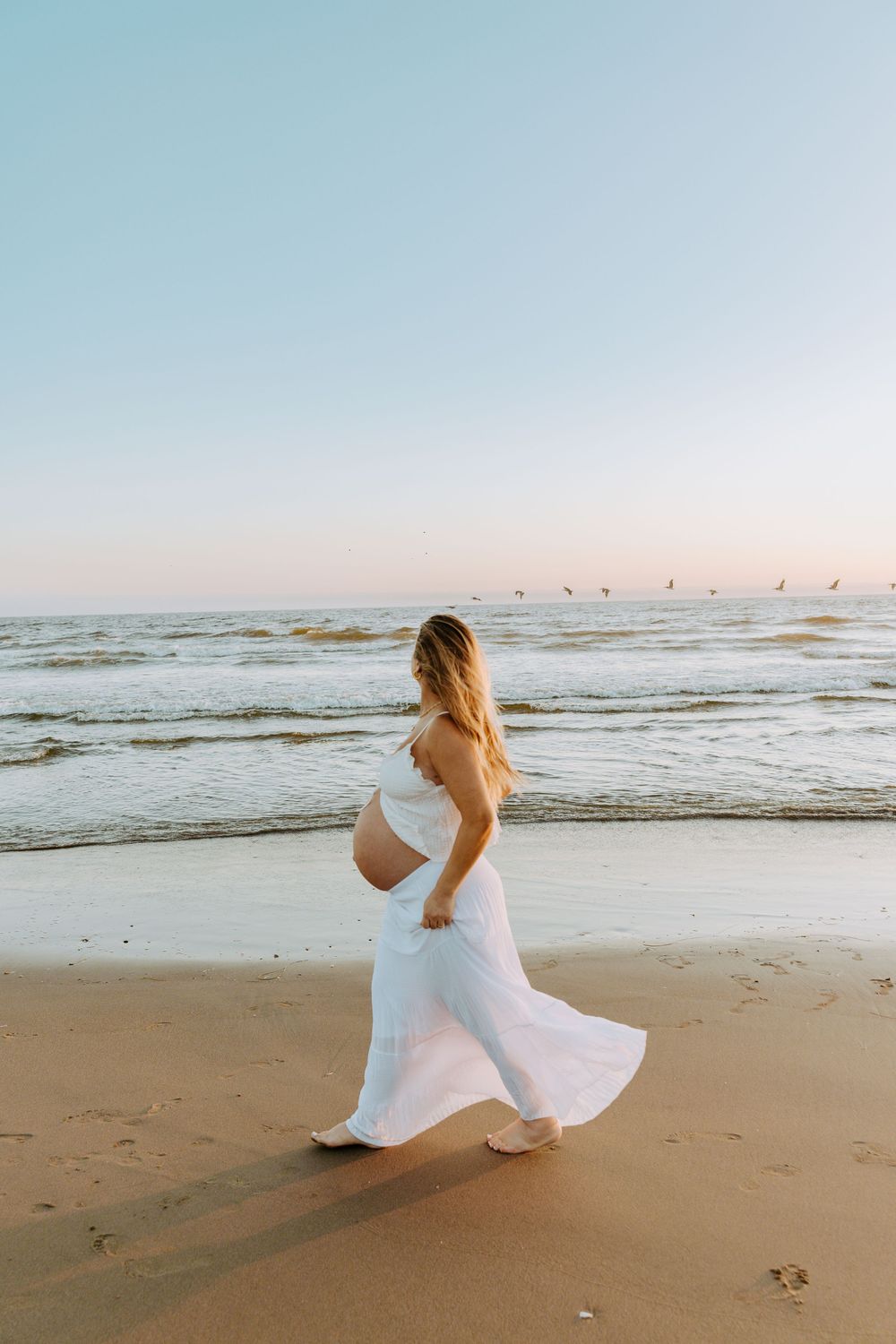 A woman in a flowing white dress walks along a beach at sunset with birds flying in the distance.
