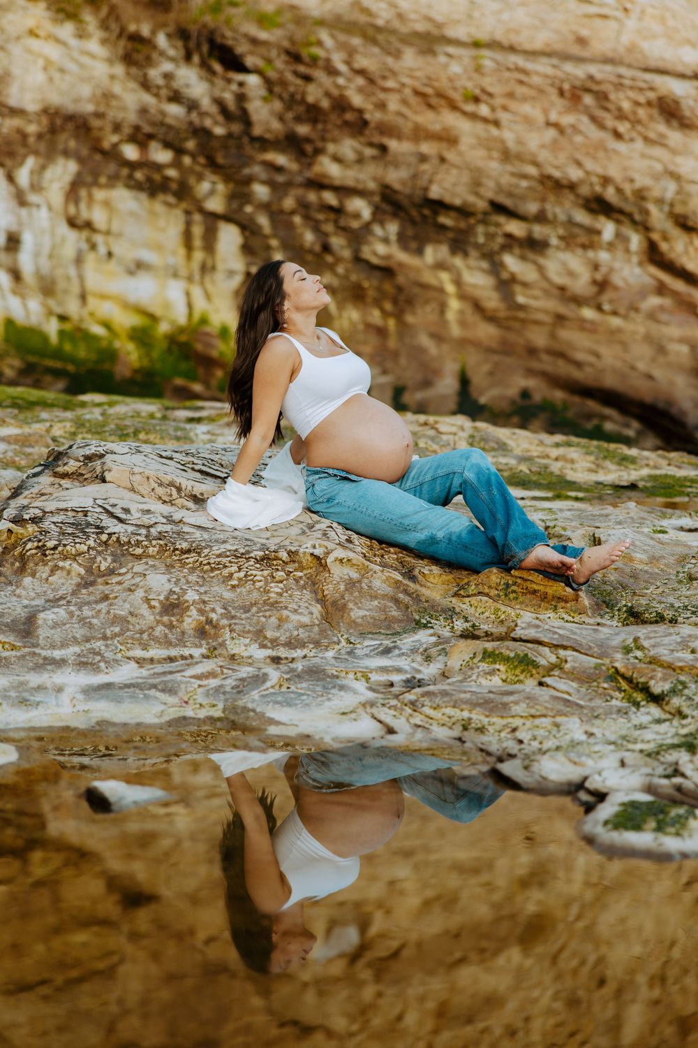 Artistic maternity portrait with reflection in tide pool against weathered coastal rock formations.