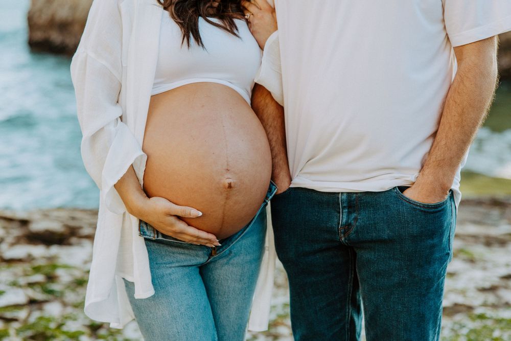 Closeup of pregnant belly with gentle hands cradling it while couple stands near water's edge.