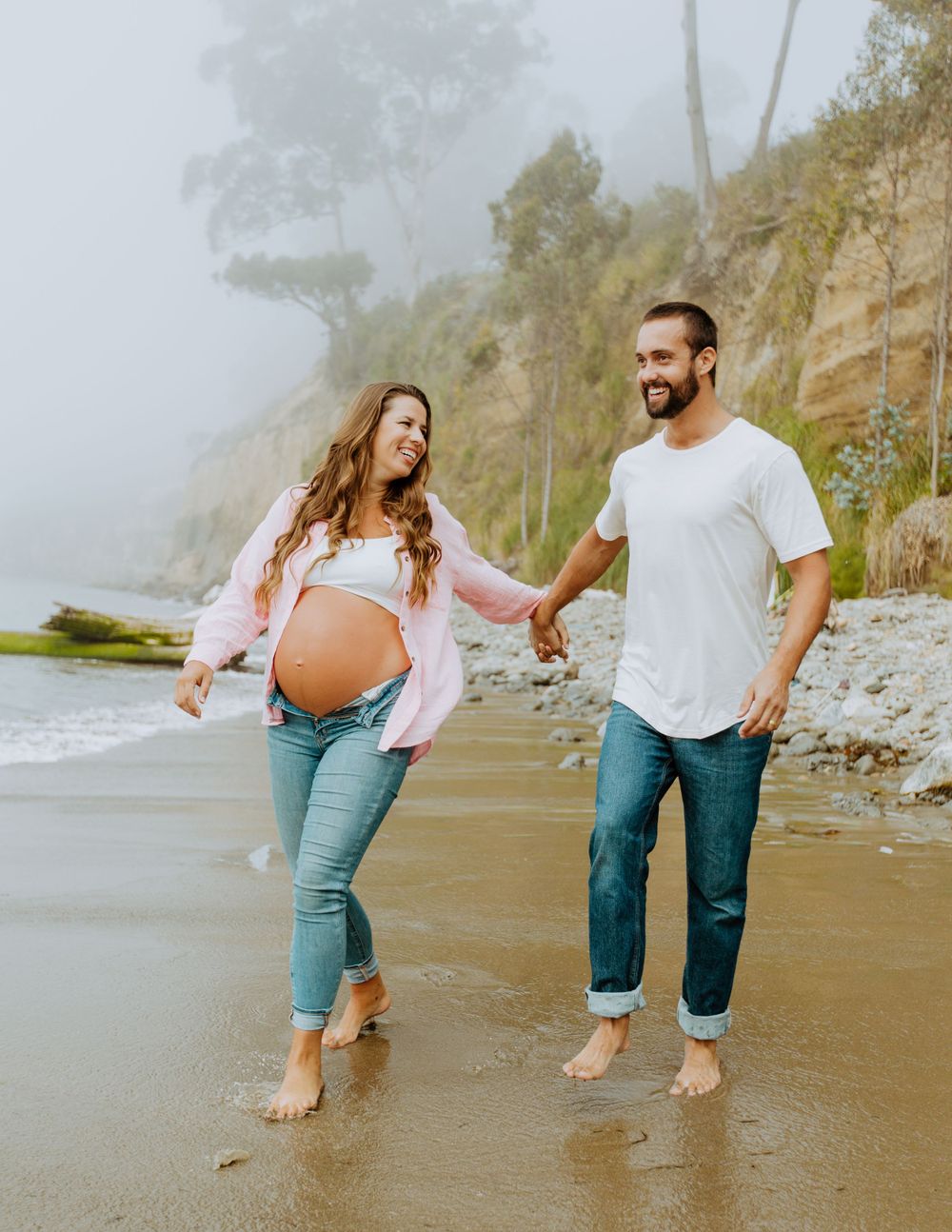 Couple walking barefoot on misty beach holding hands during maternity photoshoot.
