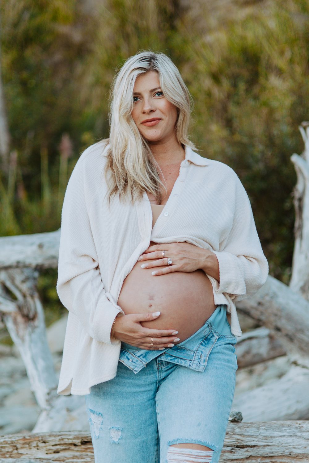A maternity photoshoot on a beach with driftwood features a white button-down shirt and distressed light blue jeans.