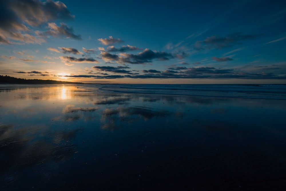 Dramatic sunset reflects on calm ocean waters with dark clouds and vibrant blue sky during golden hour.