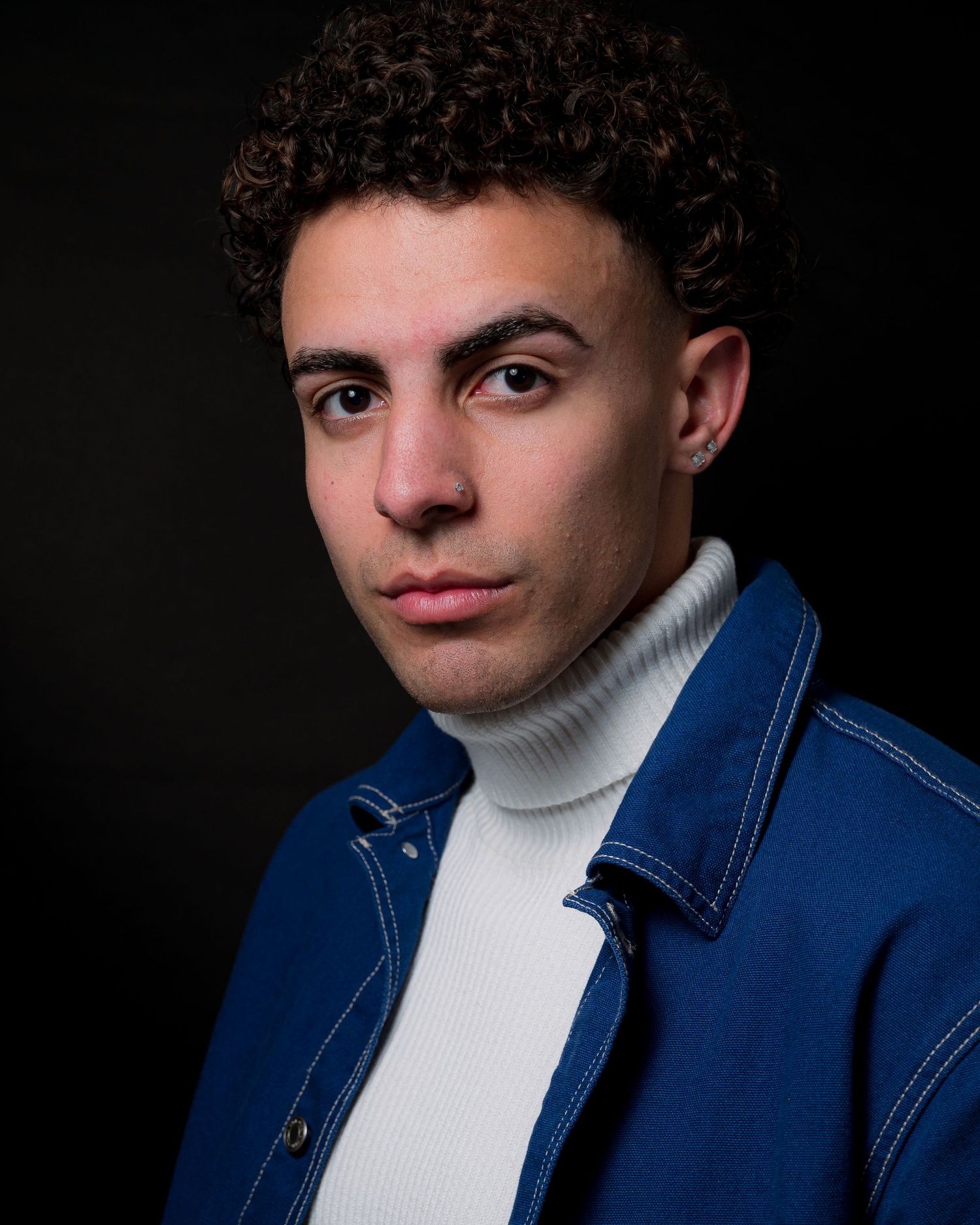 Stylish portrait in white turtleneck and blue denim jacket against dark background.