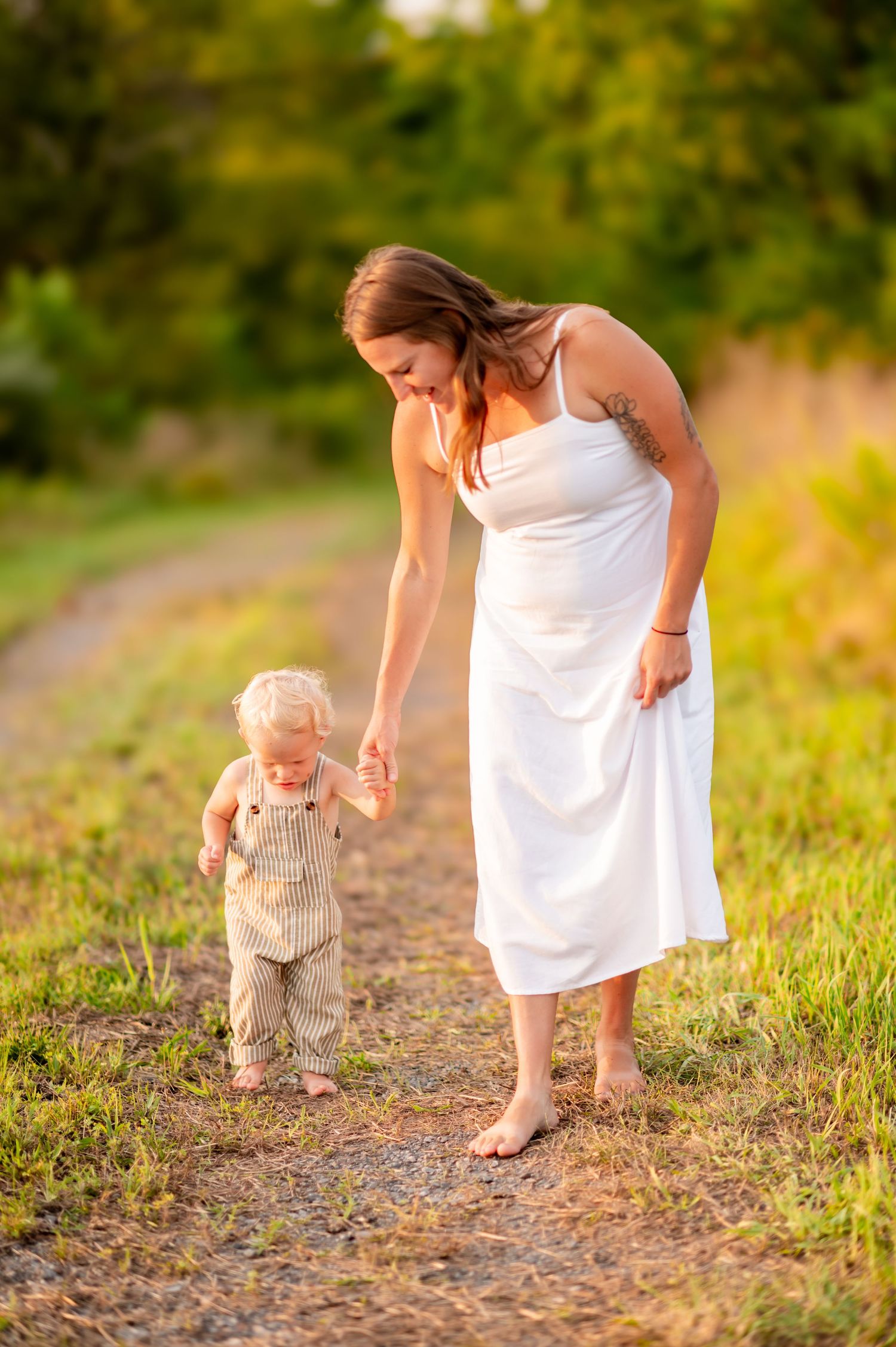 A parent and toddler in overalls walking together on a sunlit dirt path during golden hour.