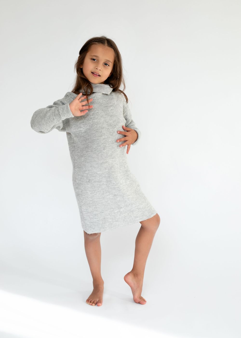 Dancer in gray sweater dress poses playfully against white studio background.