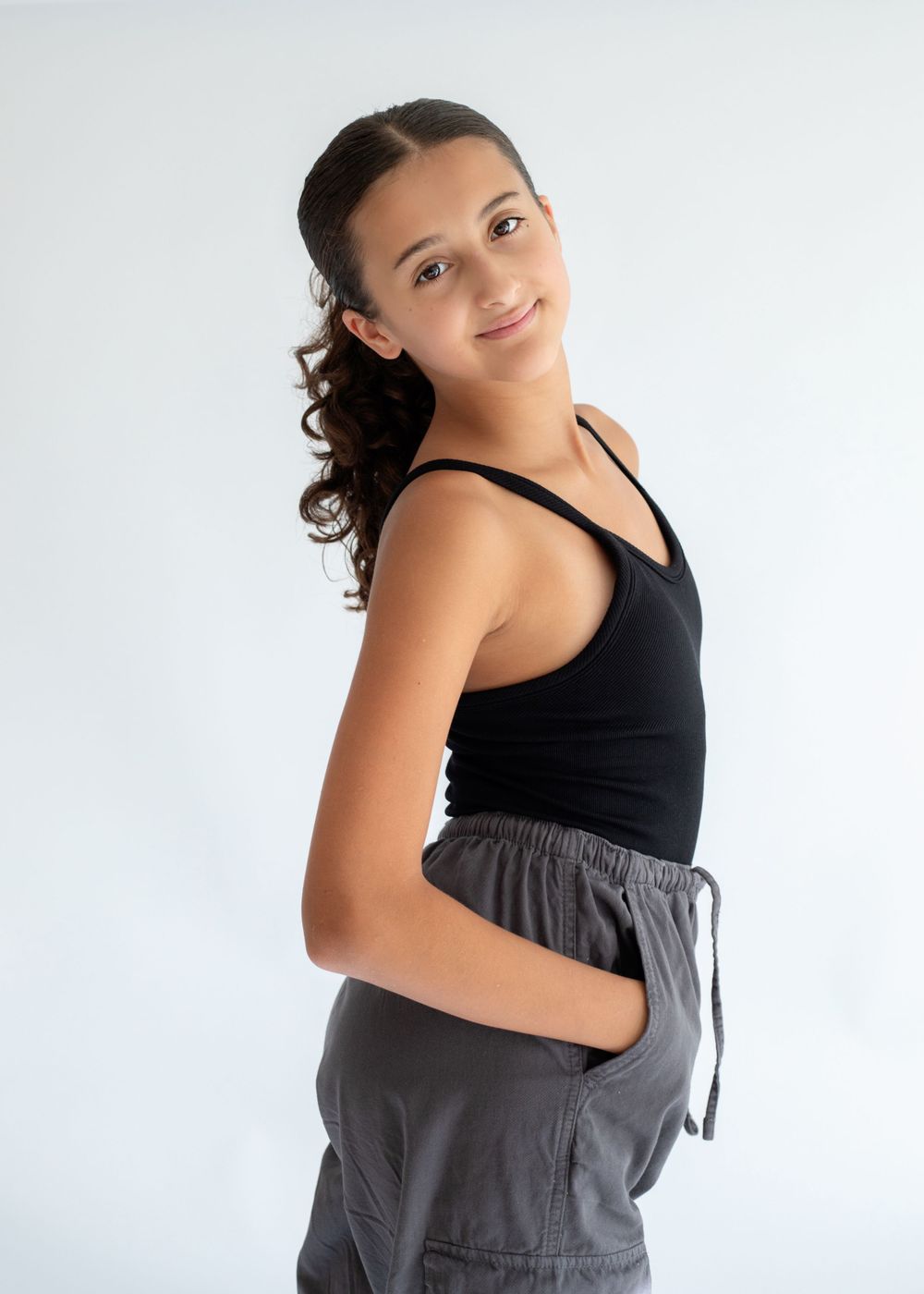 Young dancer in black tank top and gray pants smiles while posing in studio setting.