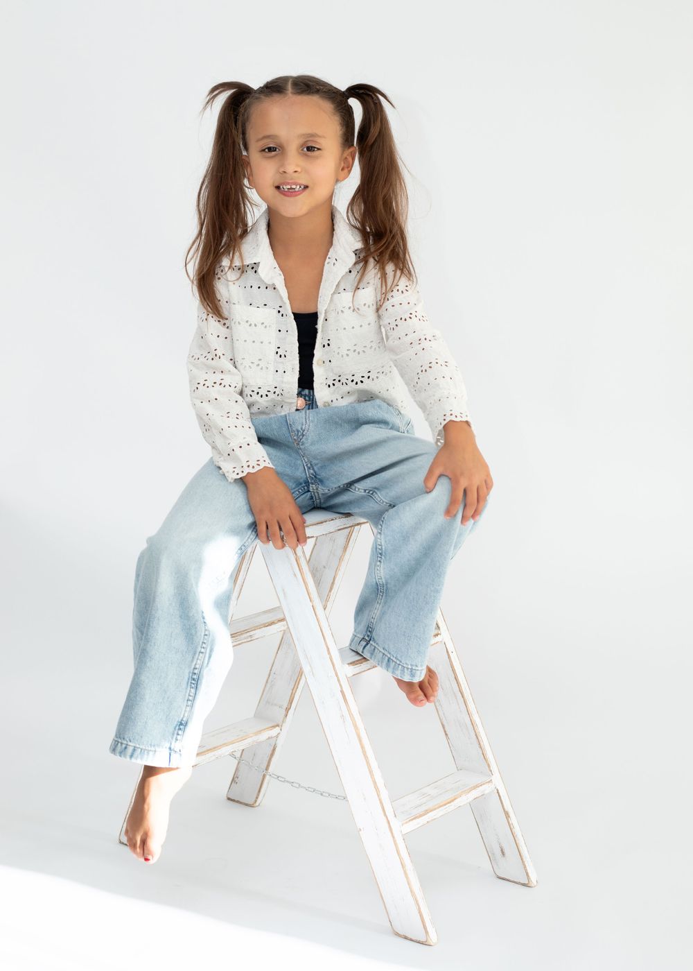A young model in a white crochet cardigan and light jeans poses casually on a white stepladder.
