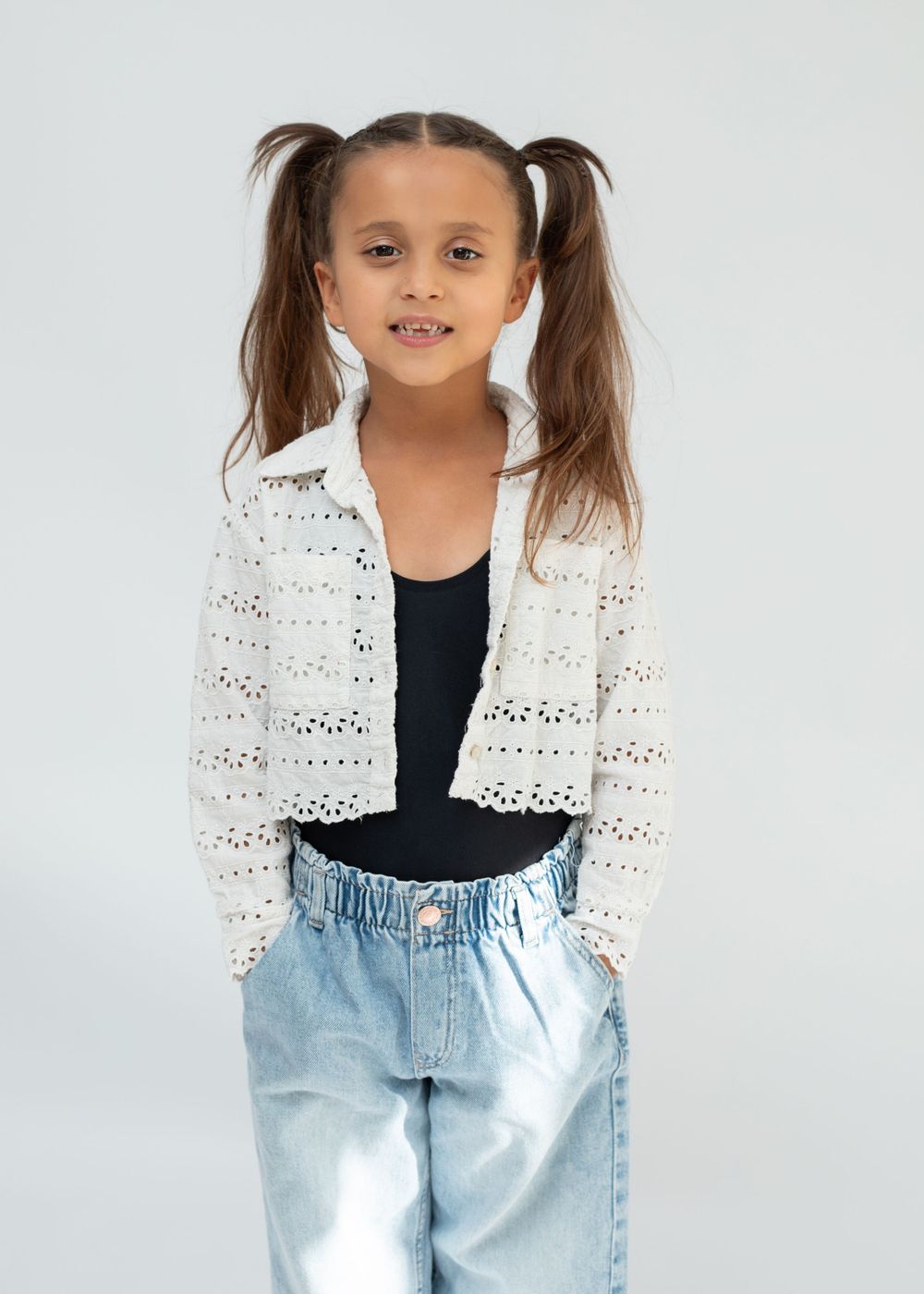 Model wearing white crochet cardigan and jeans poses against clean white backdrop.