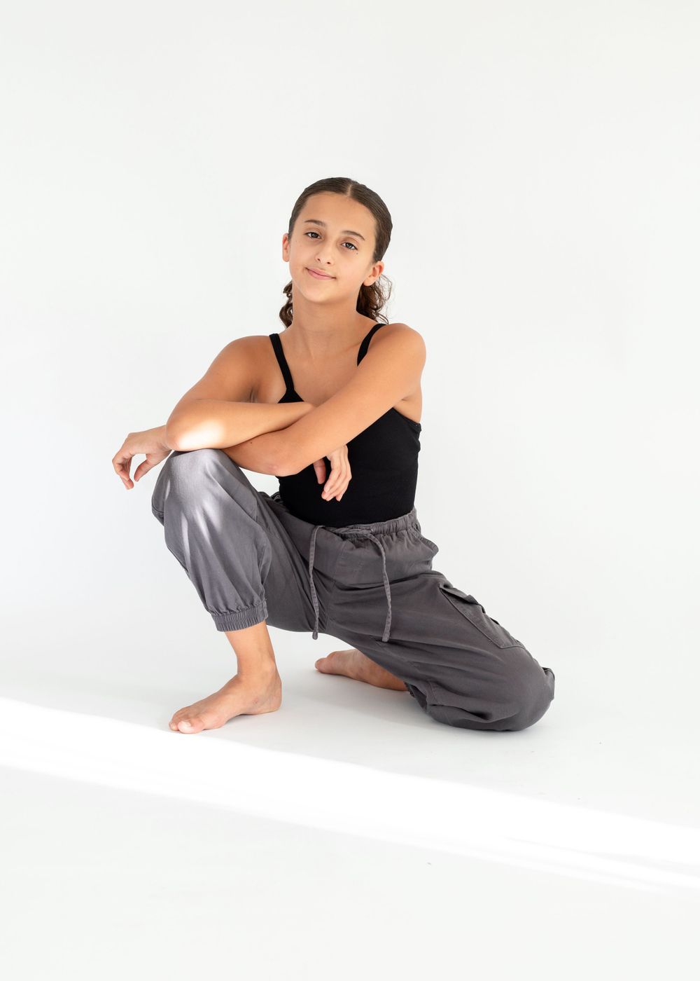 Athletic dancer in black tank top and gray joggers poses in a crouched position against white background.